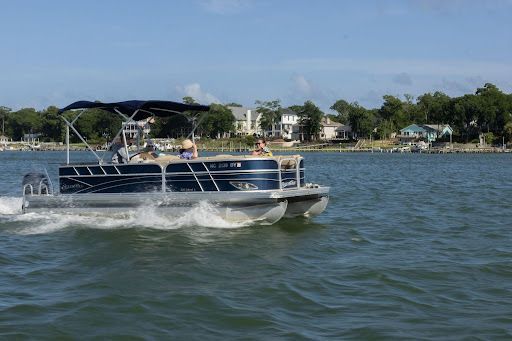 Pontoon boat cruising on a lake with water spray and shoreline houses in the background