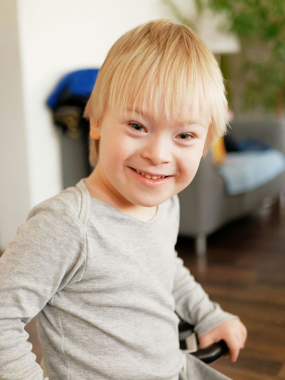 A young boy with down syndrome is smiling for the camera while sitting in a wheelchair.