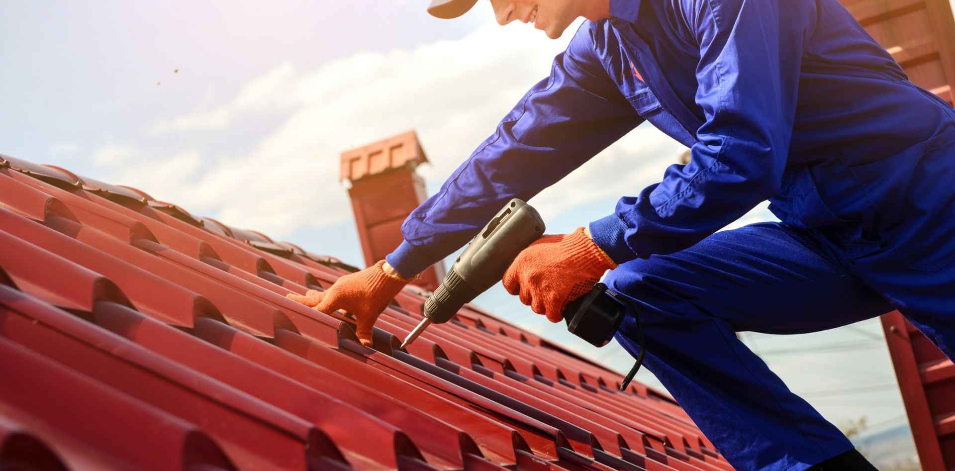 Roofer in blue jumpsuit using a drill on a red metal roof.