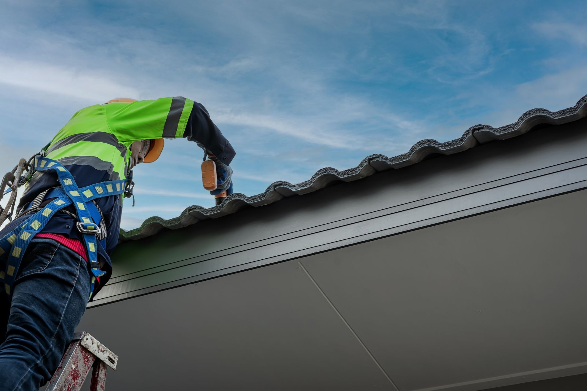 Roofer in safety gear, drilling into a corrugated metal roof under a blue sky.