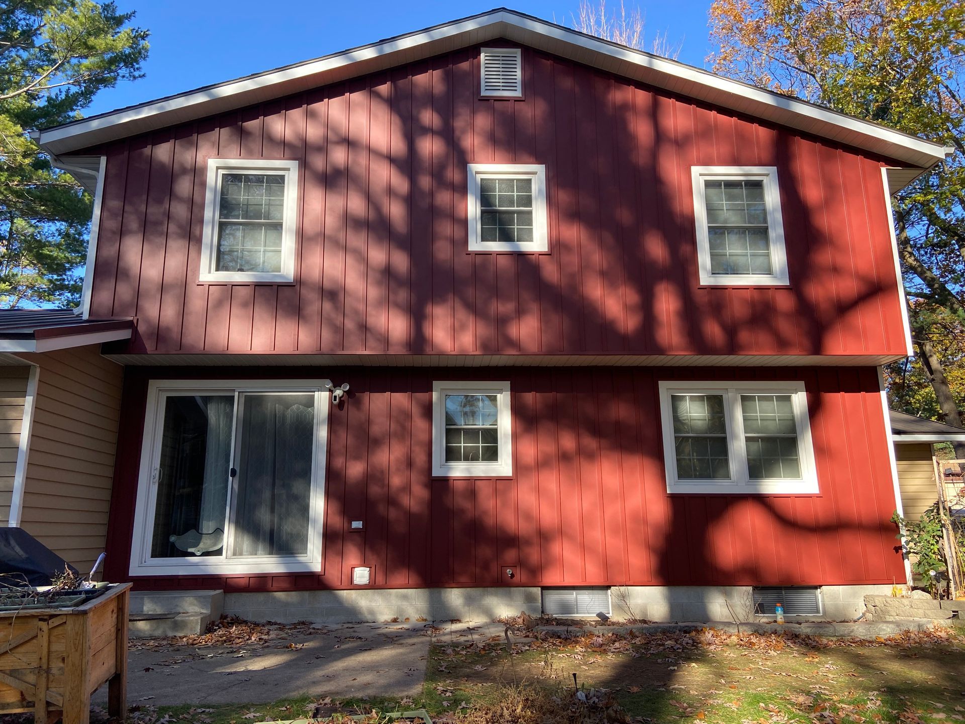 Red two-story house with white-trimmed windows and sliding glass door. Shadows of trees cast across the siding.