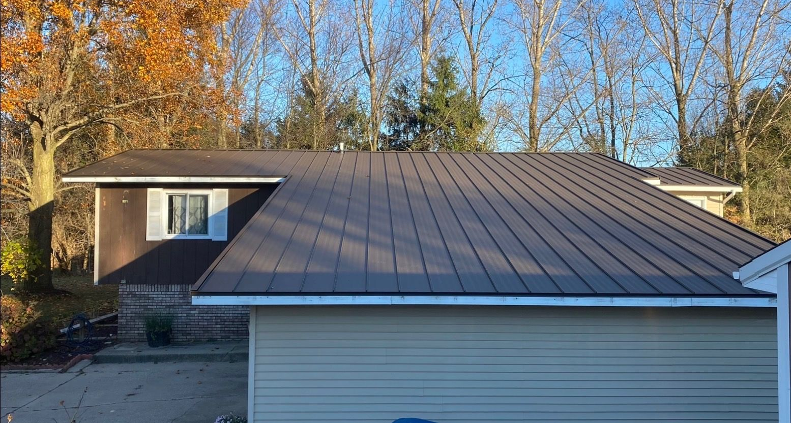 Brown metal roof on a house with beige siding and a garage. Trees and blue sky in the background.