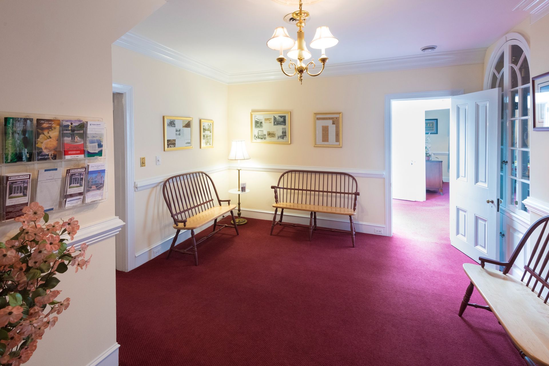 Bright waiting room with red carpet, wooden benches, chandelier, and open white doors.
