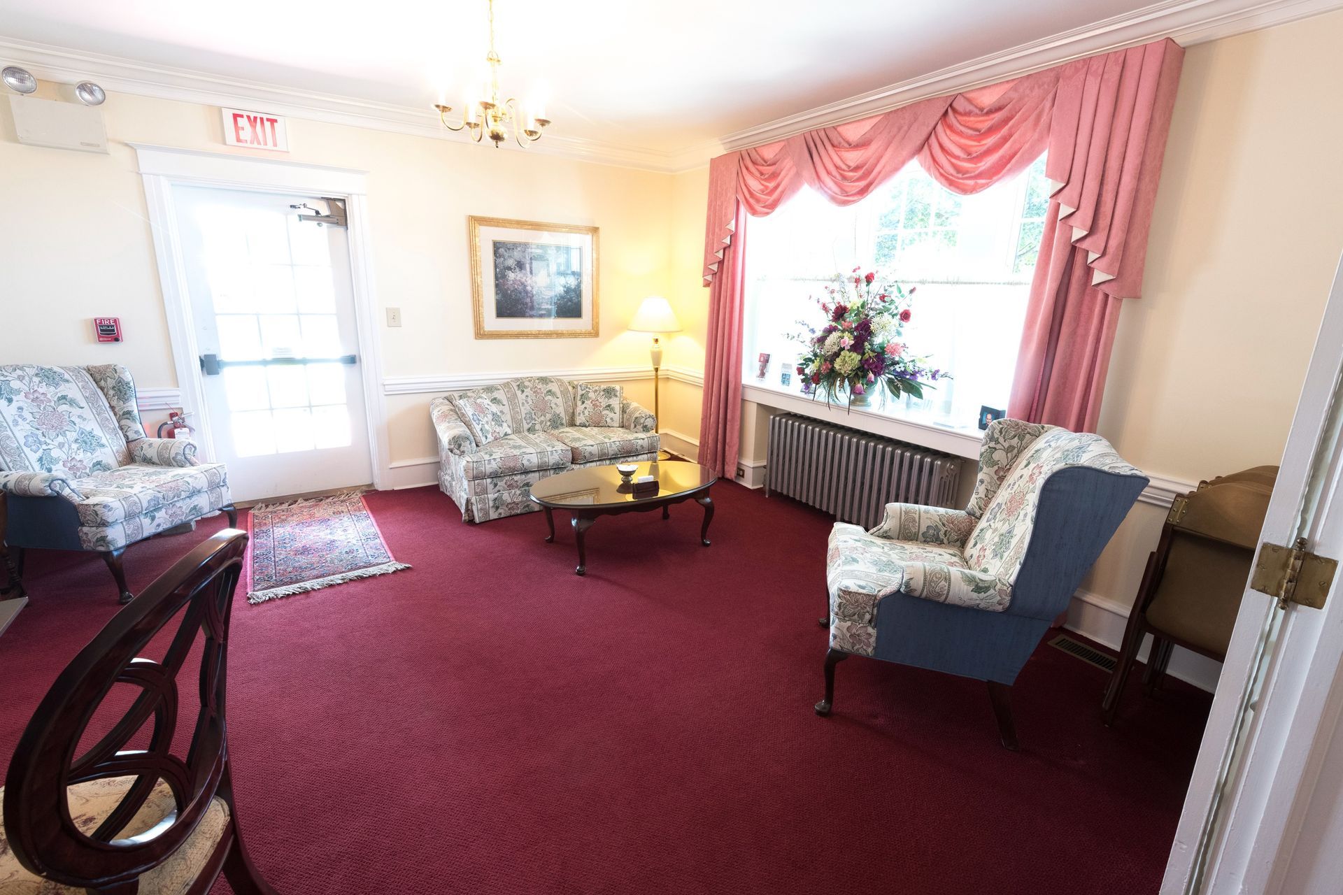 Bright living room with burgundy carpet, floral sofas, blue armchairs, and a window with pink drapes