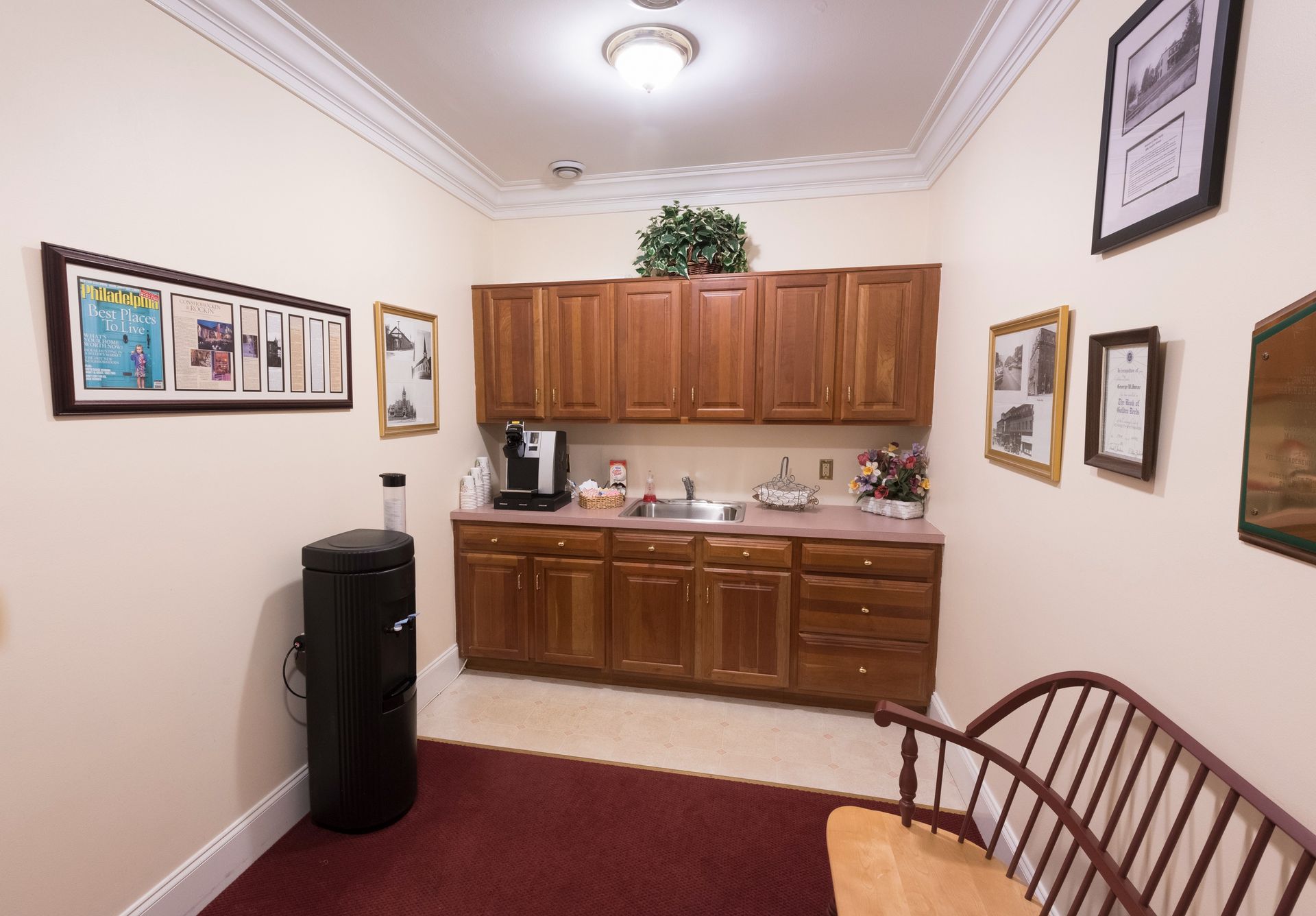 Small waiting room with wooden cabinets, coffee maker, framed wall art, and a red rug.