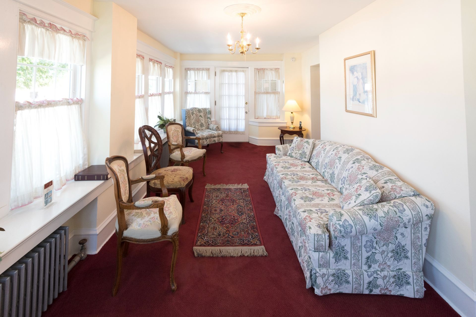Bright sunroom with floral sofa, chairs, red carpet, and white curtains