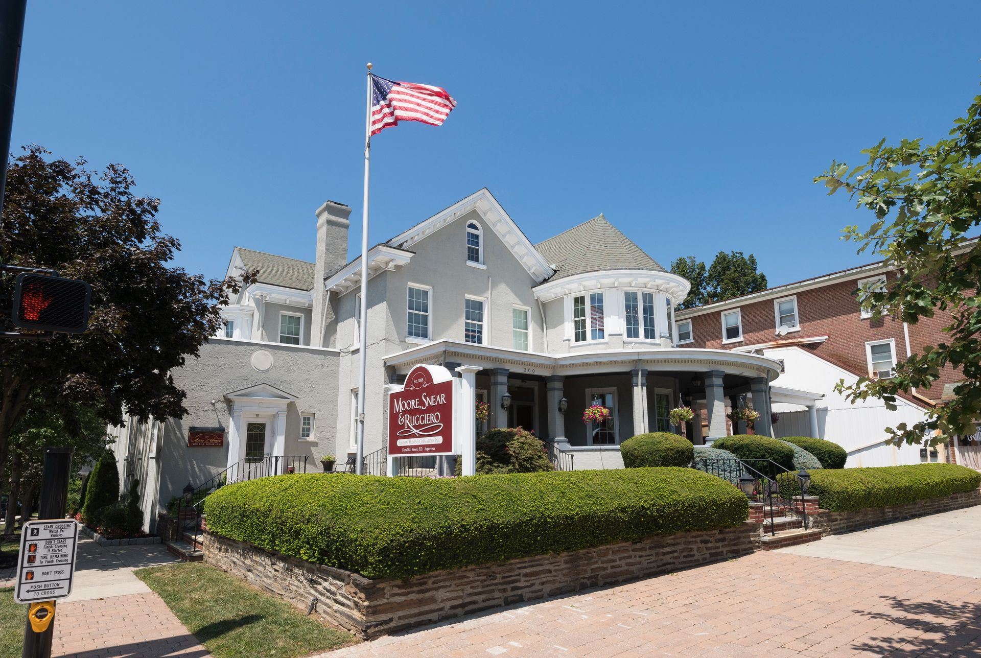 White Victorian-style house with an American flag, porch, and landscaped hedges beside a brick walkway.