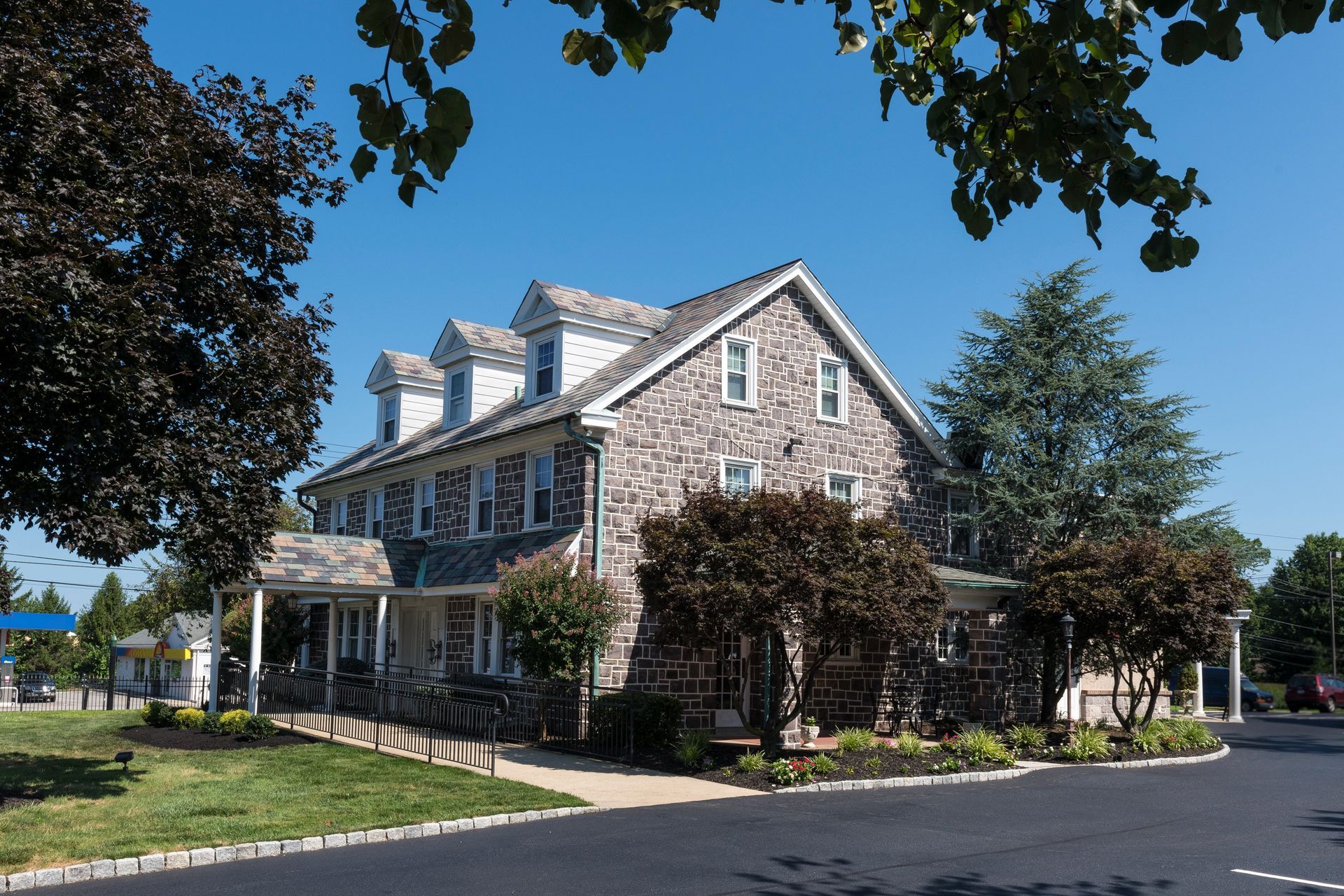 Stone house with blue-gray siding and trees, viewed from a quiet street on a sunny day