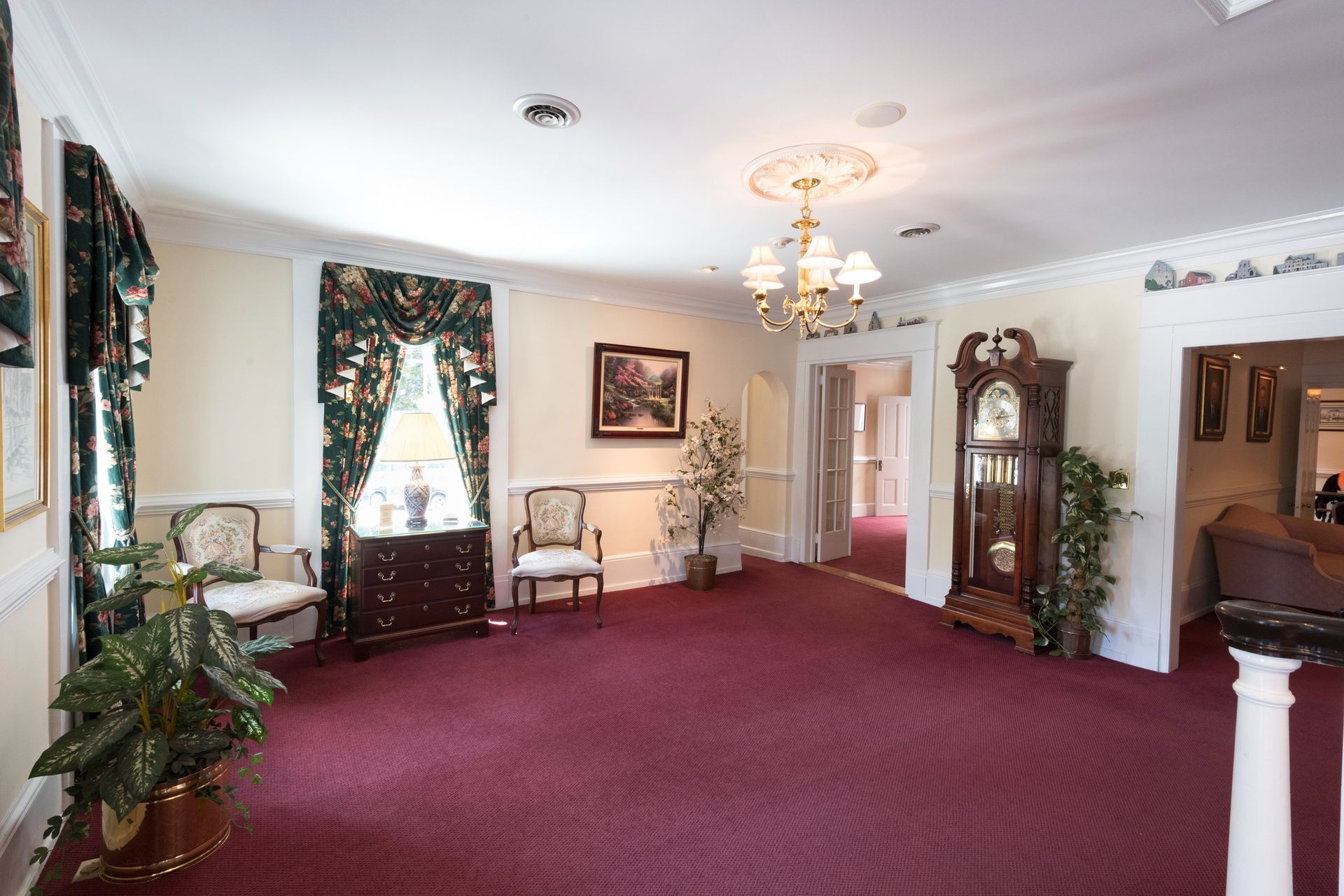 Empty carpeted formal room with floral curtains, chandelier, chairs, and a grandfather clock.