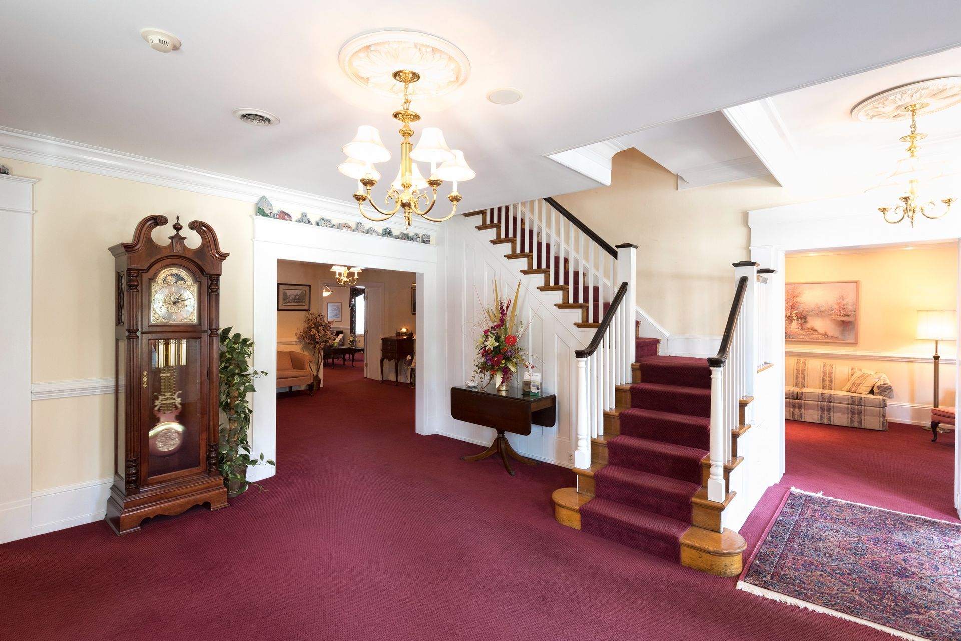 Elegant lobby with burgundy carpet, chandelier, grandfather clock, and staircase with red runner