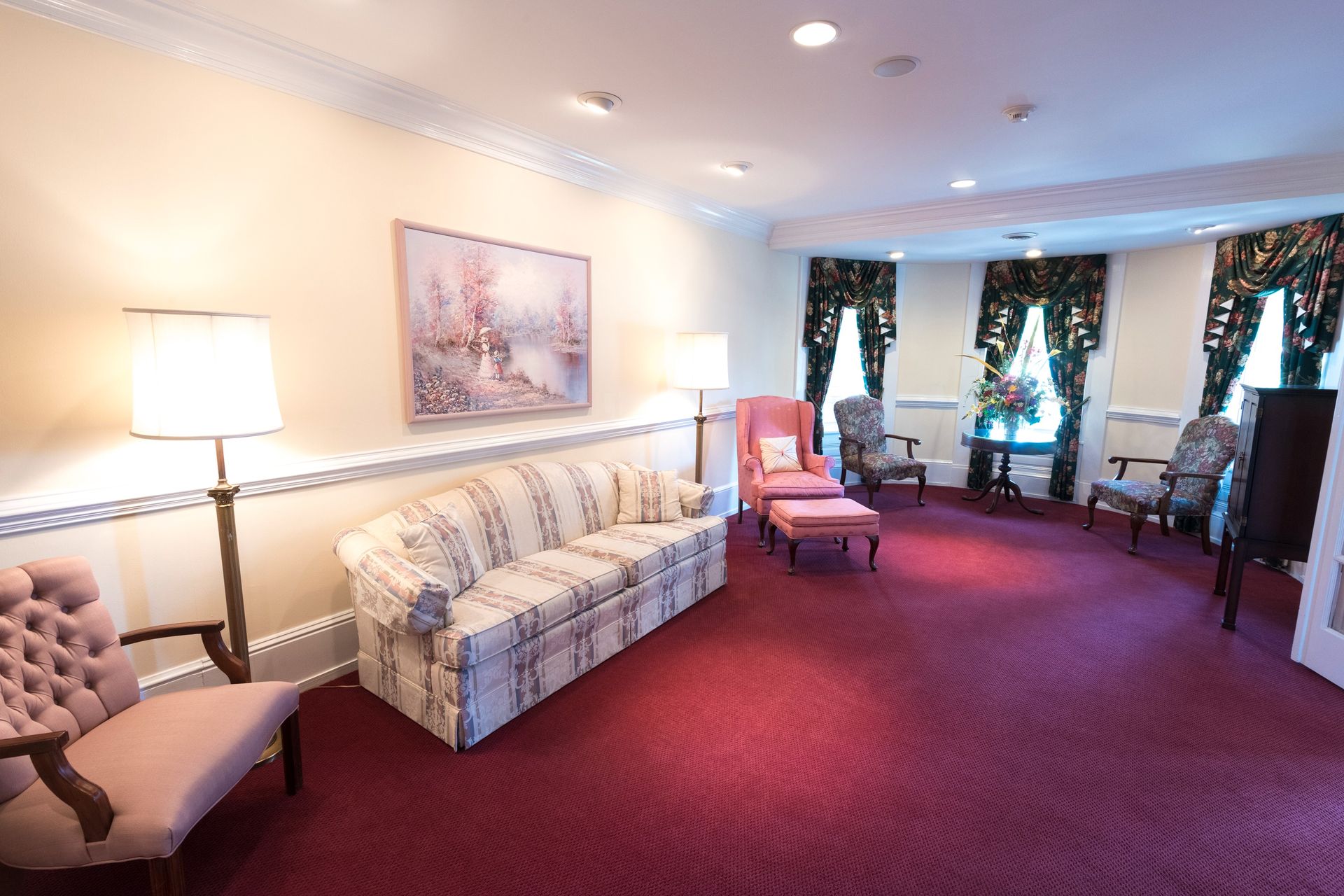 Spacious hotel lounge with patterned sofa, armchairs, lamps, and burgundy carpet by windowed wall