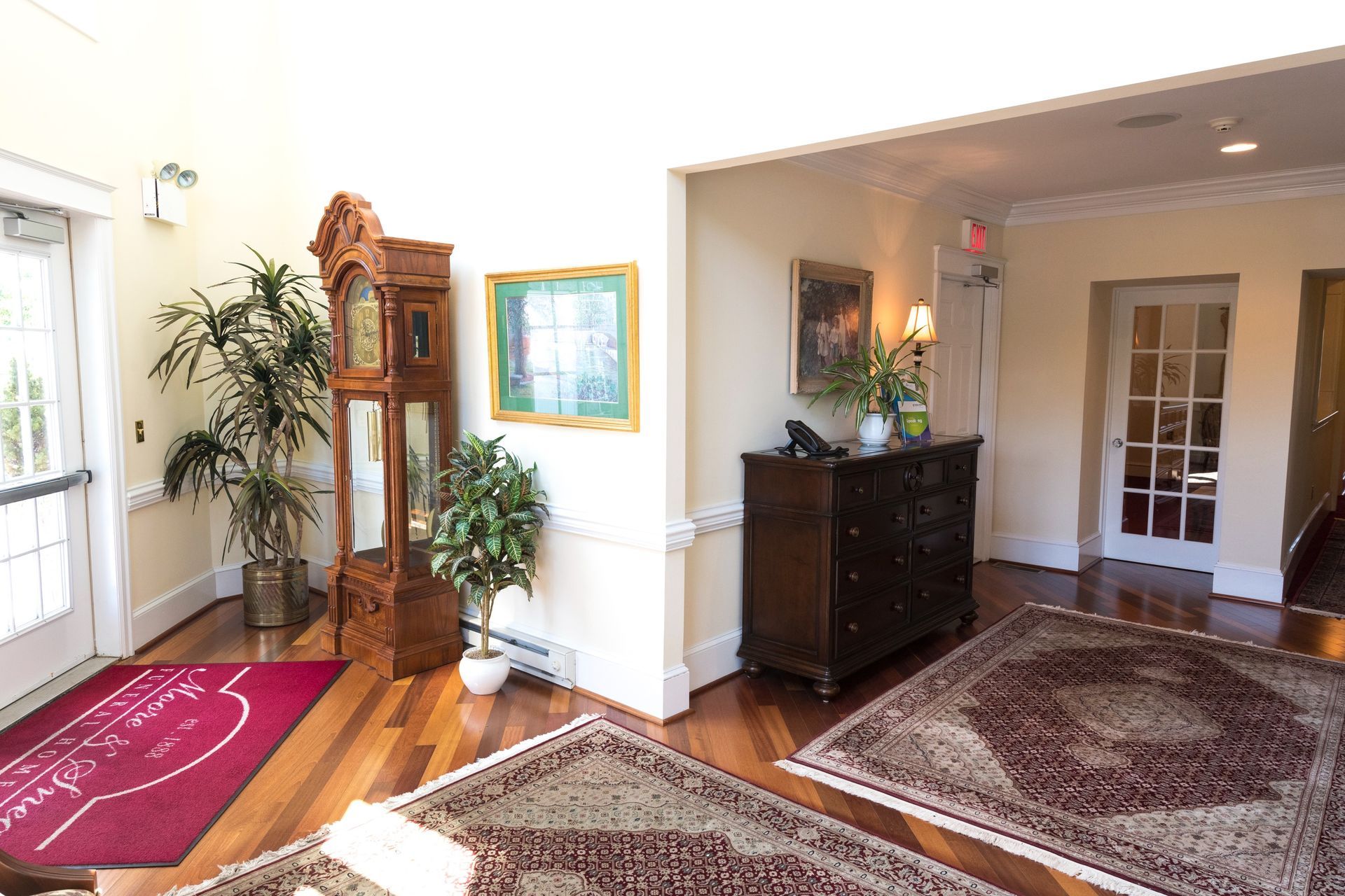 Bright foyer with rugs, plants, a wooden clock, framed art, and a dark sideboard near a glass-paneled door.