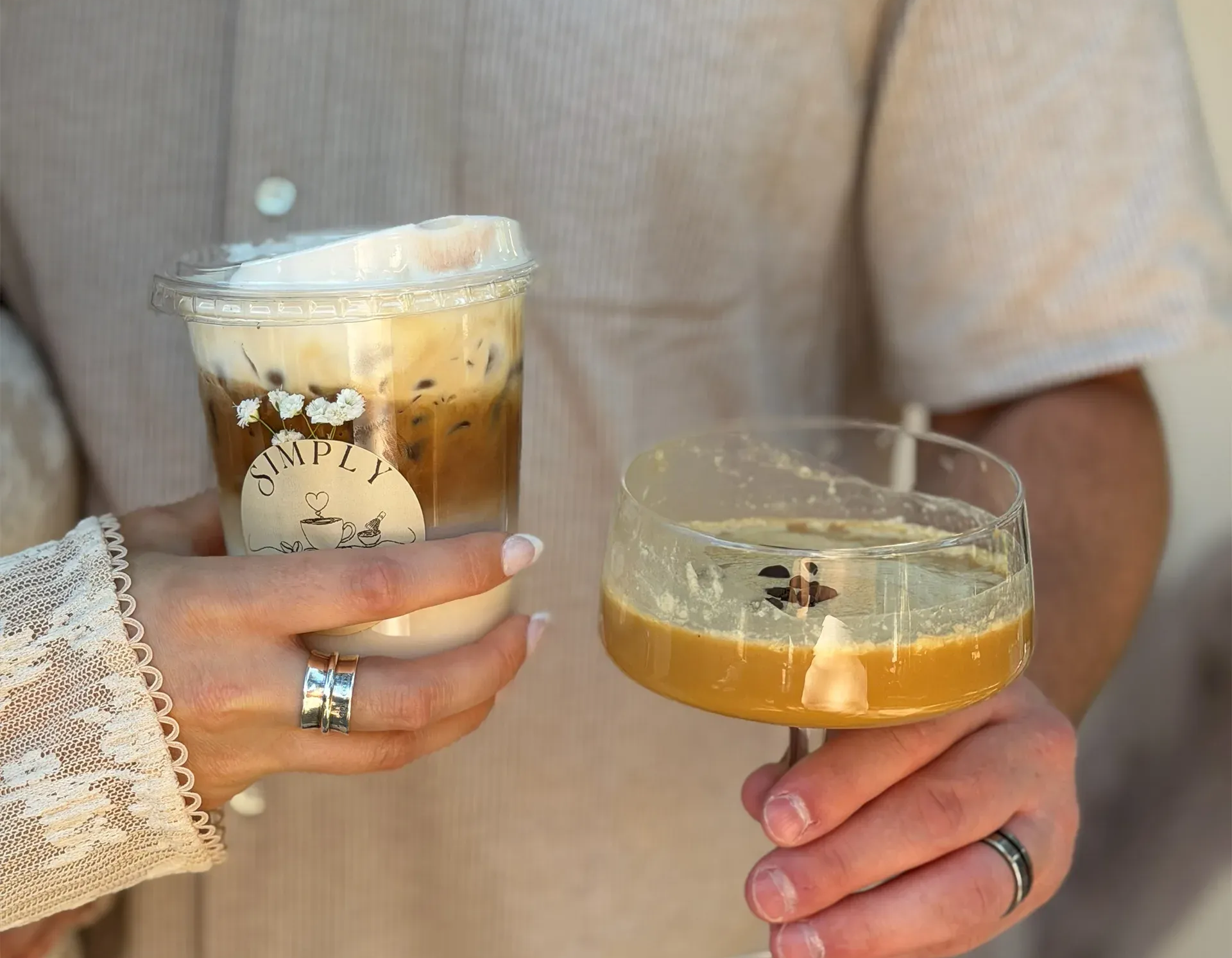 Hands holding two iced drinks, one a layered coffee and the other a clear citrus cocktail.
