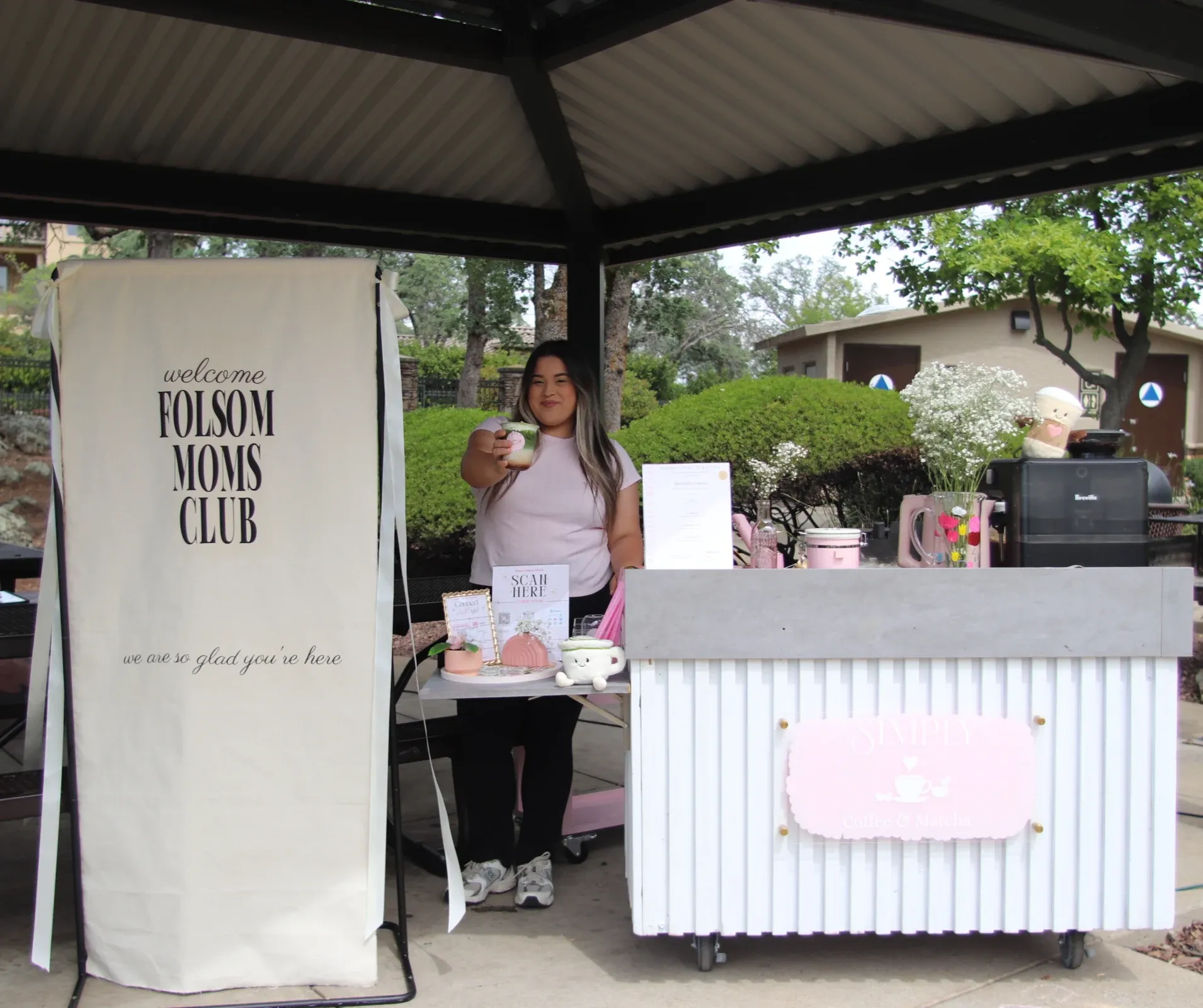 Woman at a vendor booth holding a drink beside a “Folsom Moons Club” sign and pink counter display