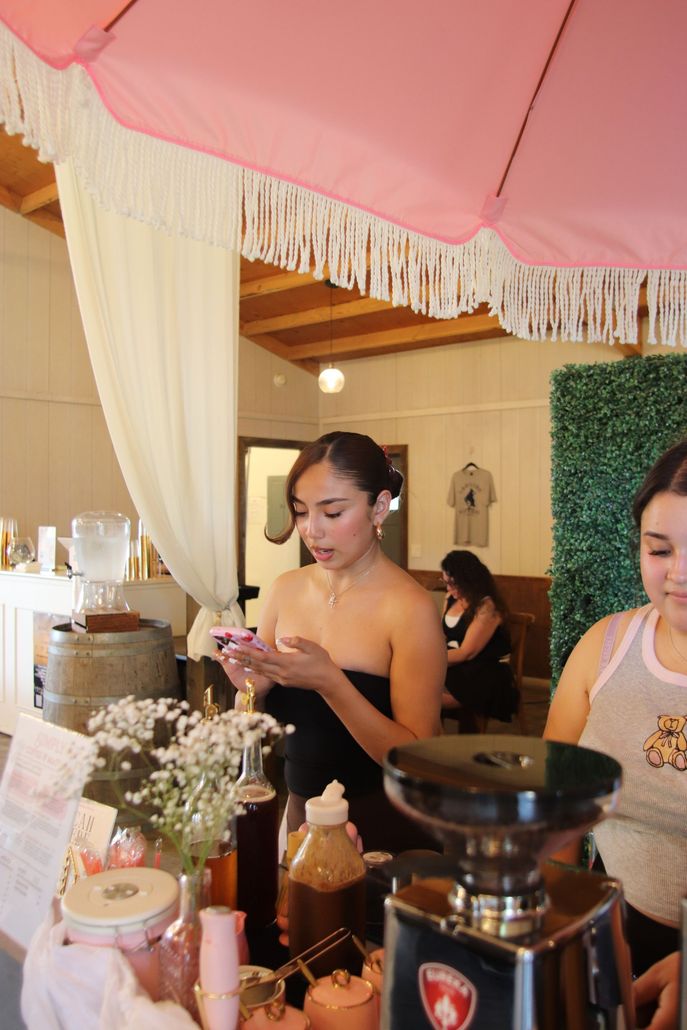 Woman behind a counter at a café, preparing a drink under a pink umbrella canopy.
