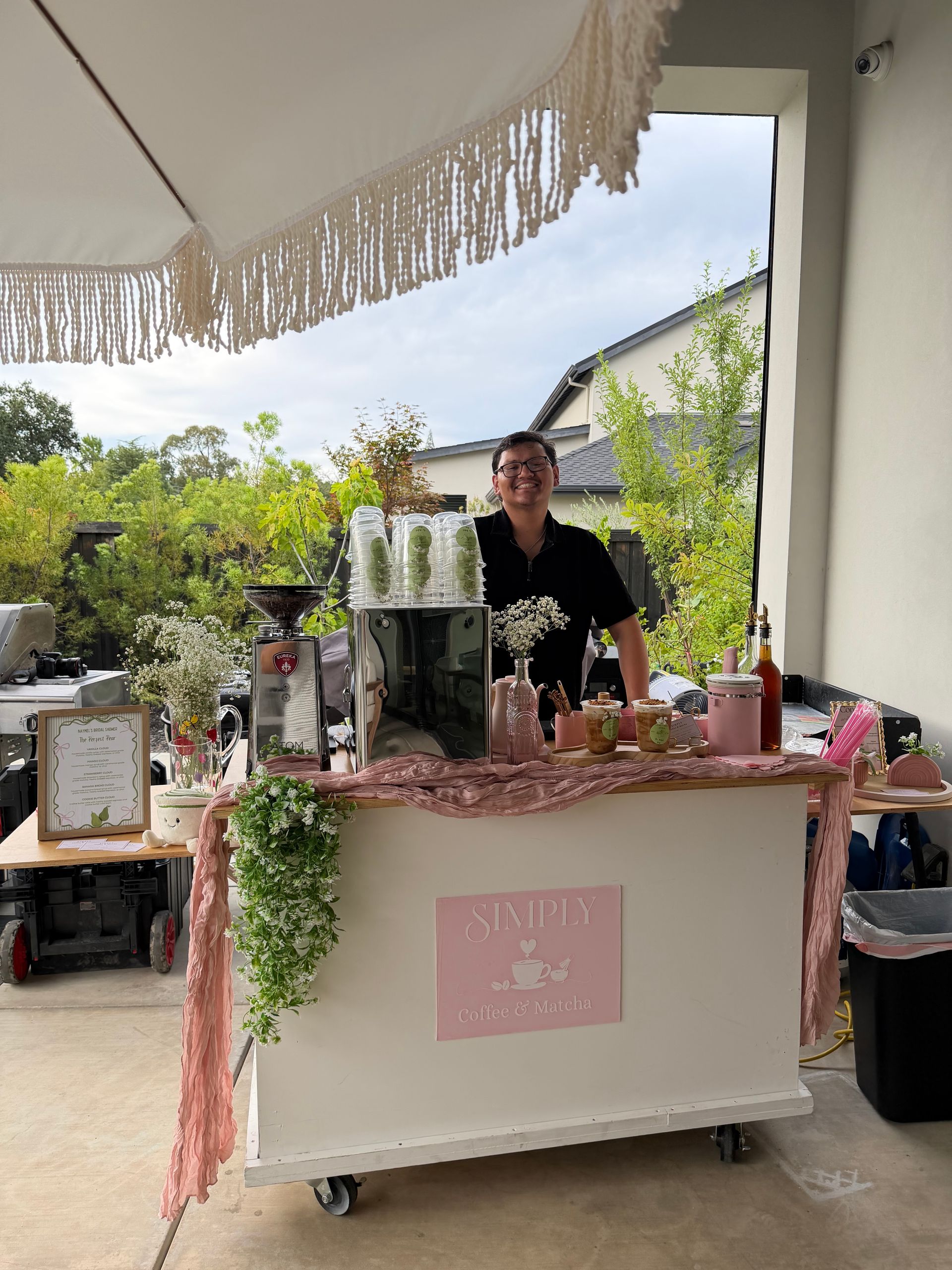 Person behind a pink drink cart with jars and bottles under a fringed umbrella outdoors