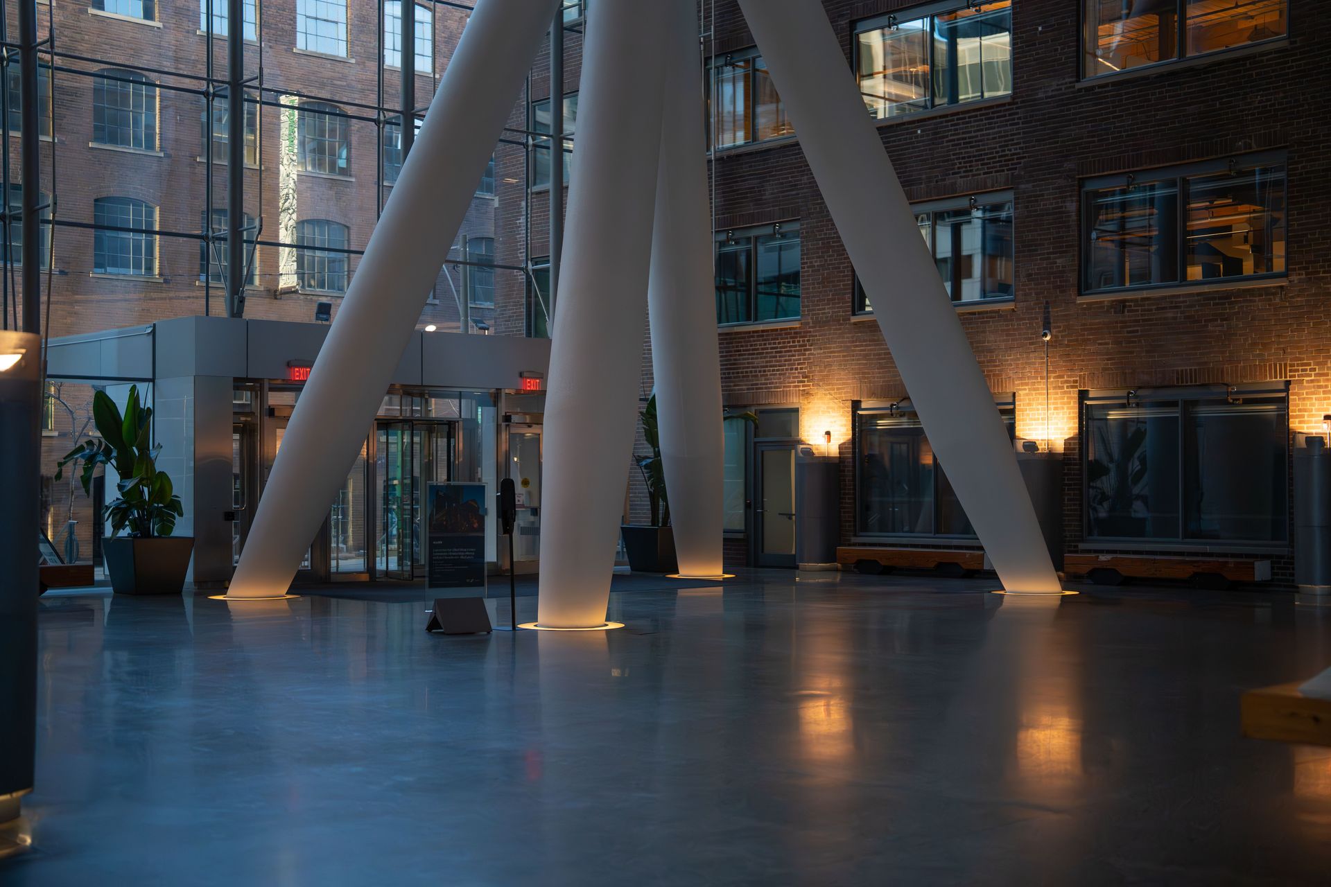 Modern indoor atrium with tall white columns, brick walls, and reflections on a polished floor at dusk