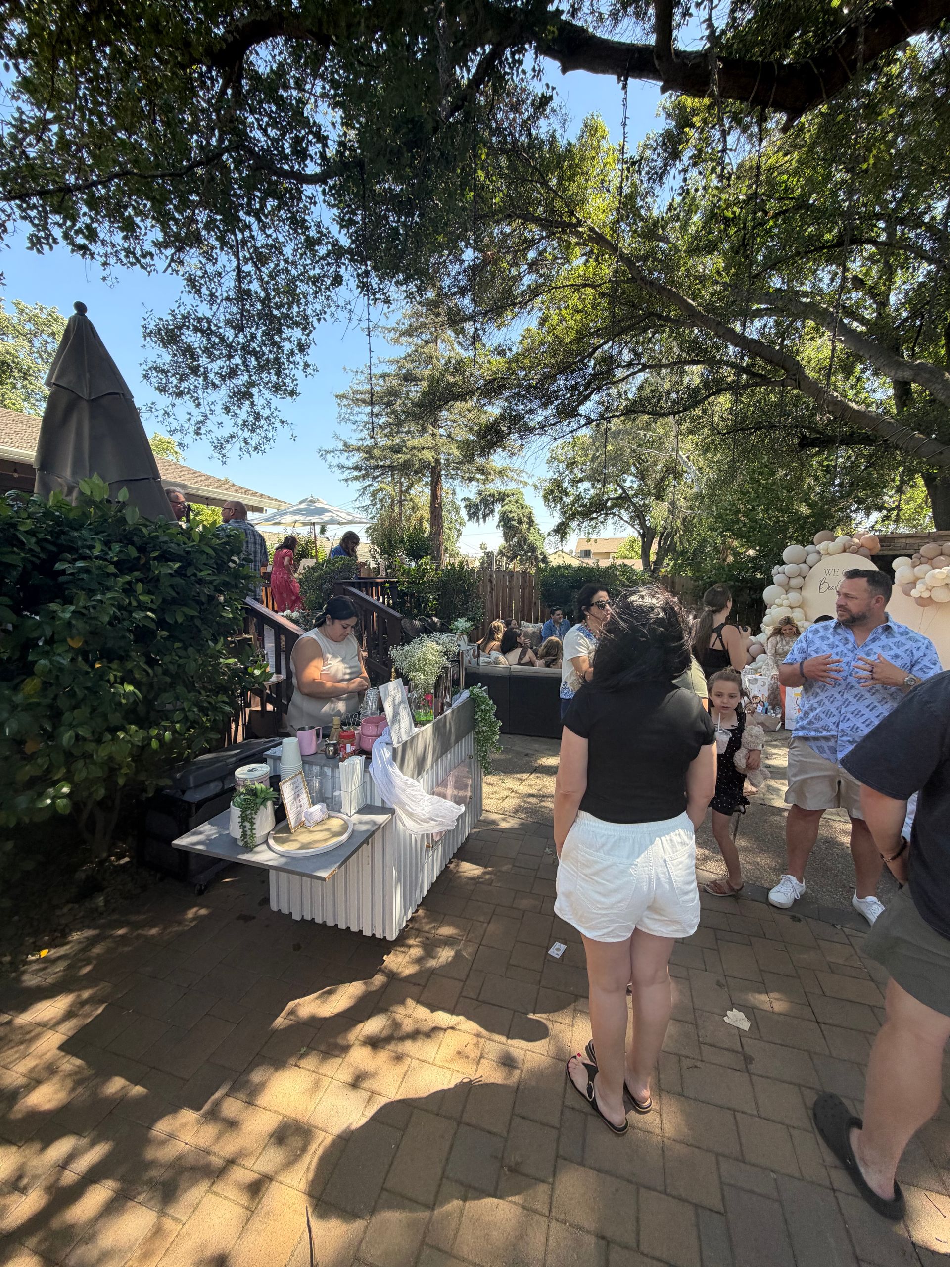 People socializing outdoors at a patio table with drinks and snacks under trees in warm sunlight