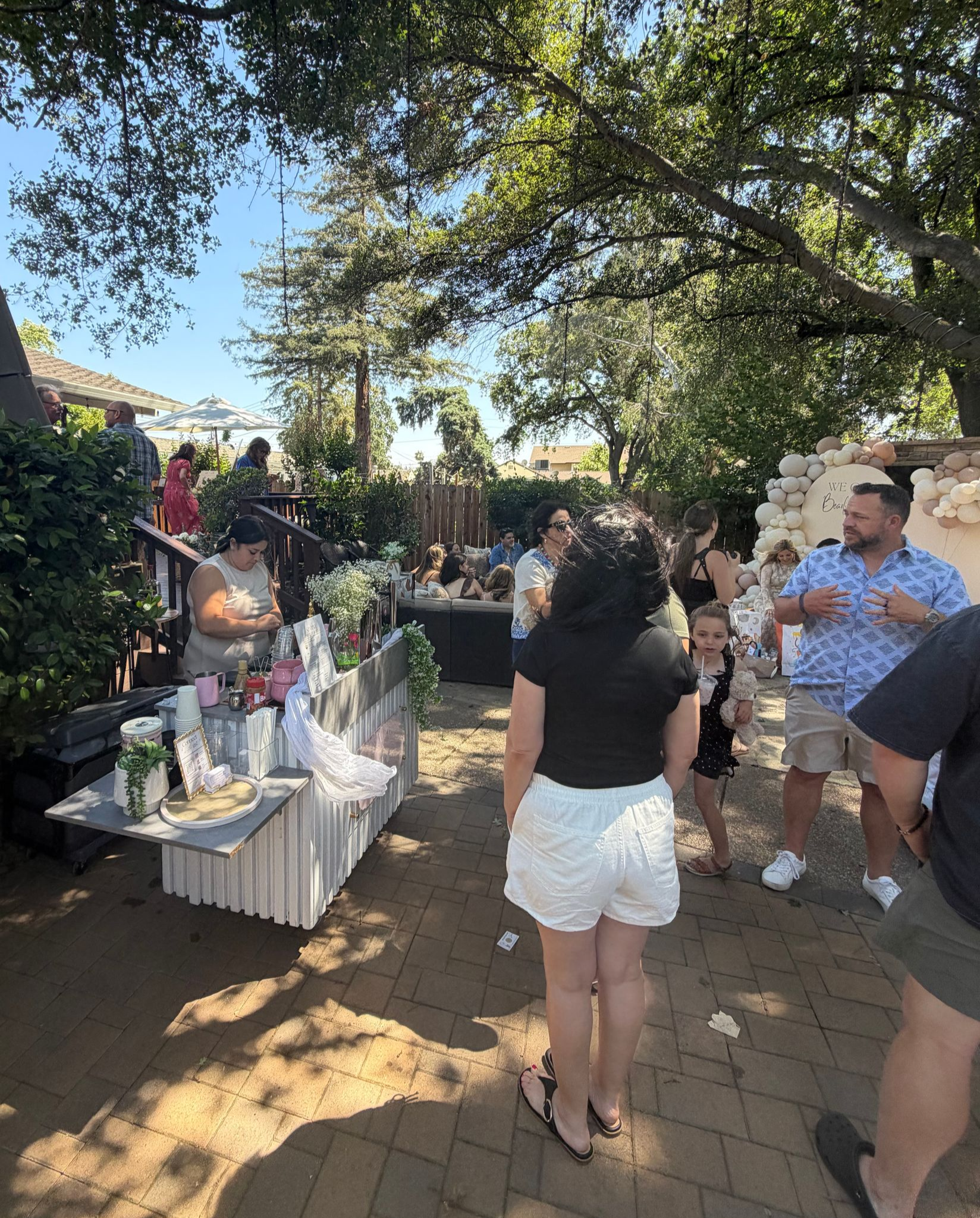 Guests mingle at an outdoor garden gathering beside a dessert table under trees.