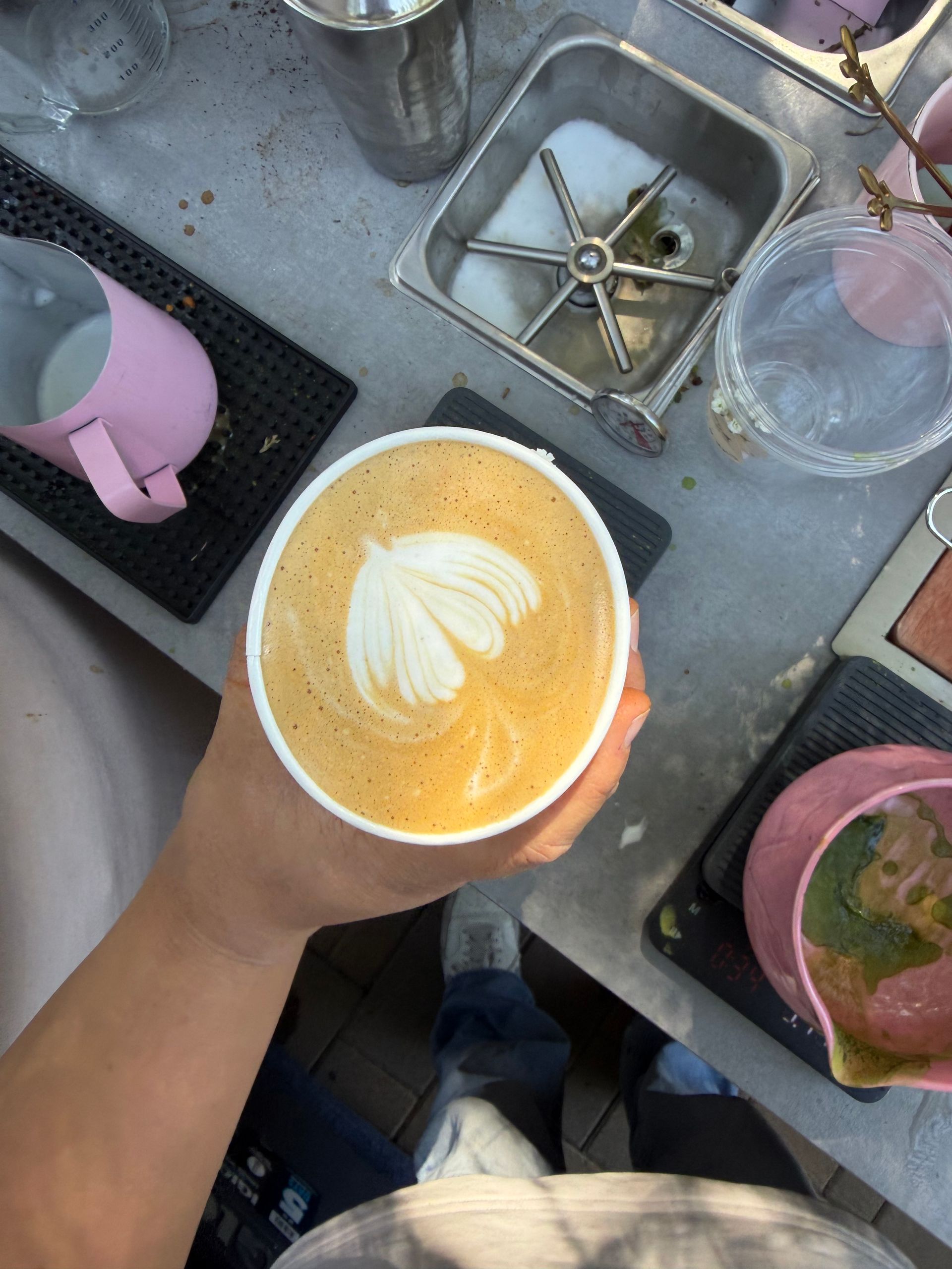 Top-down view of a hand holding a latte with leaf latte art beside other drinks on a cafe counter.
