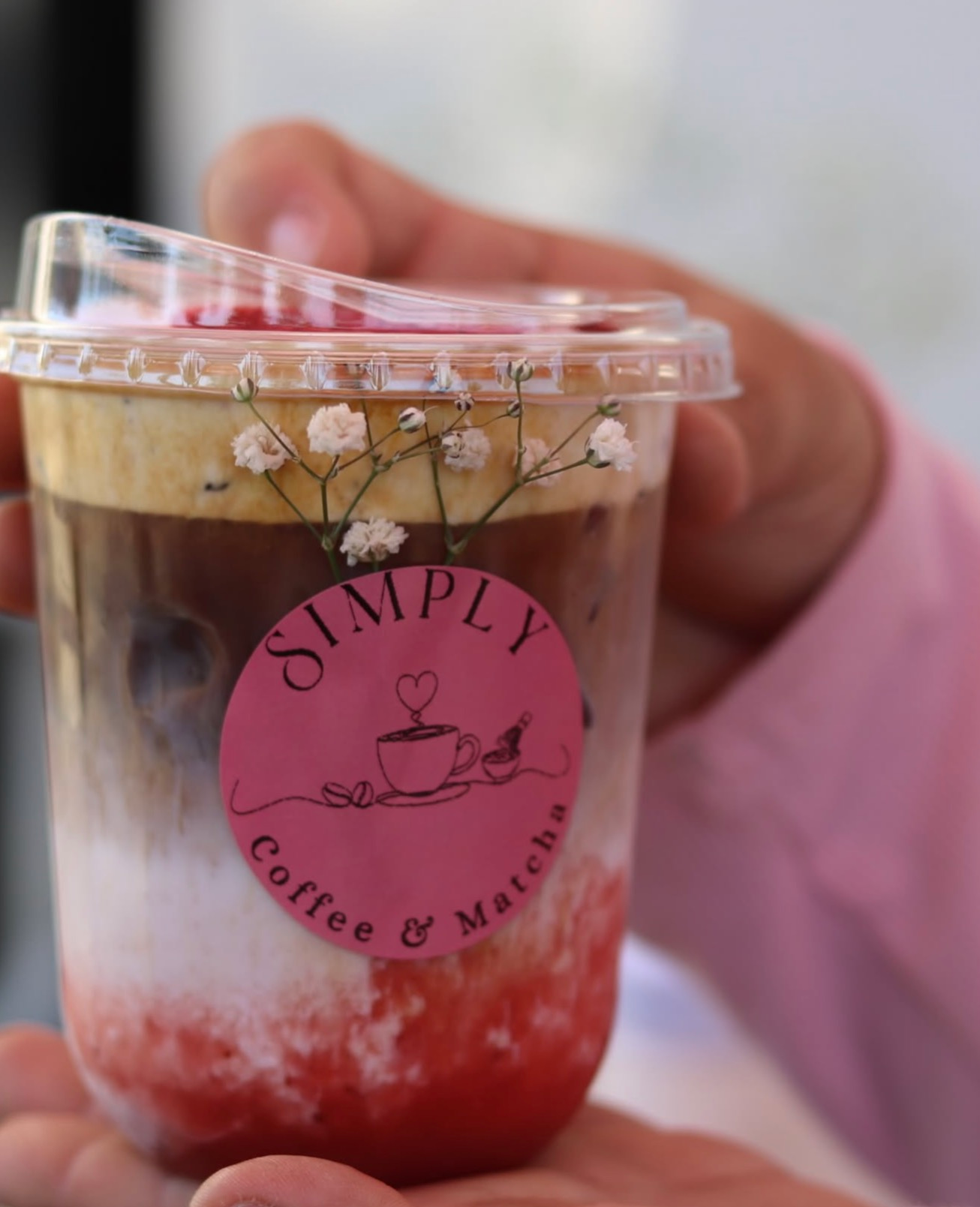 Hand holding a clear cup of layered iced coffee with a pink “Simply Coffee & More” logo