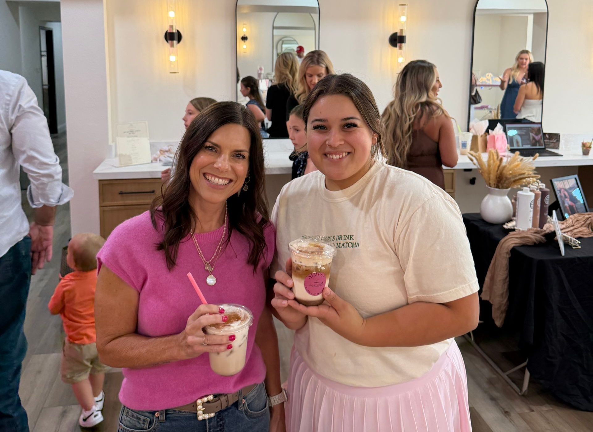 Two women smiling and holding drinks in a bright salon, with mirrored stations in the background