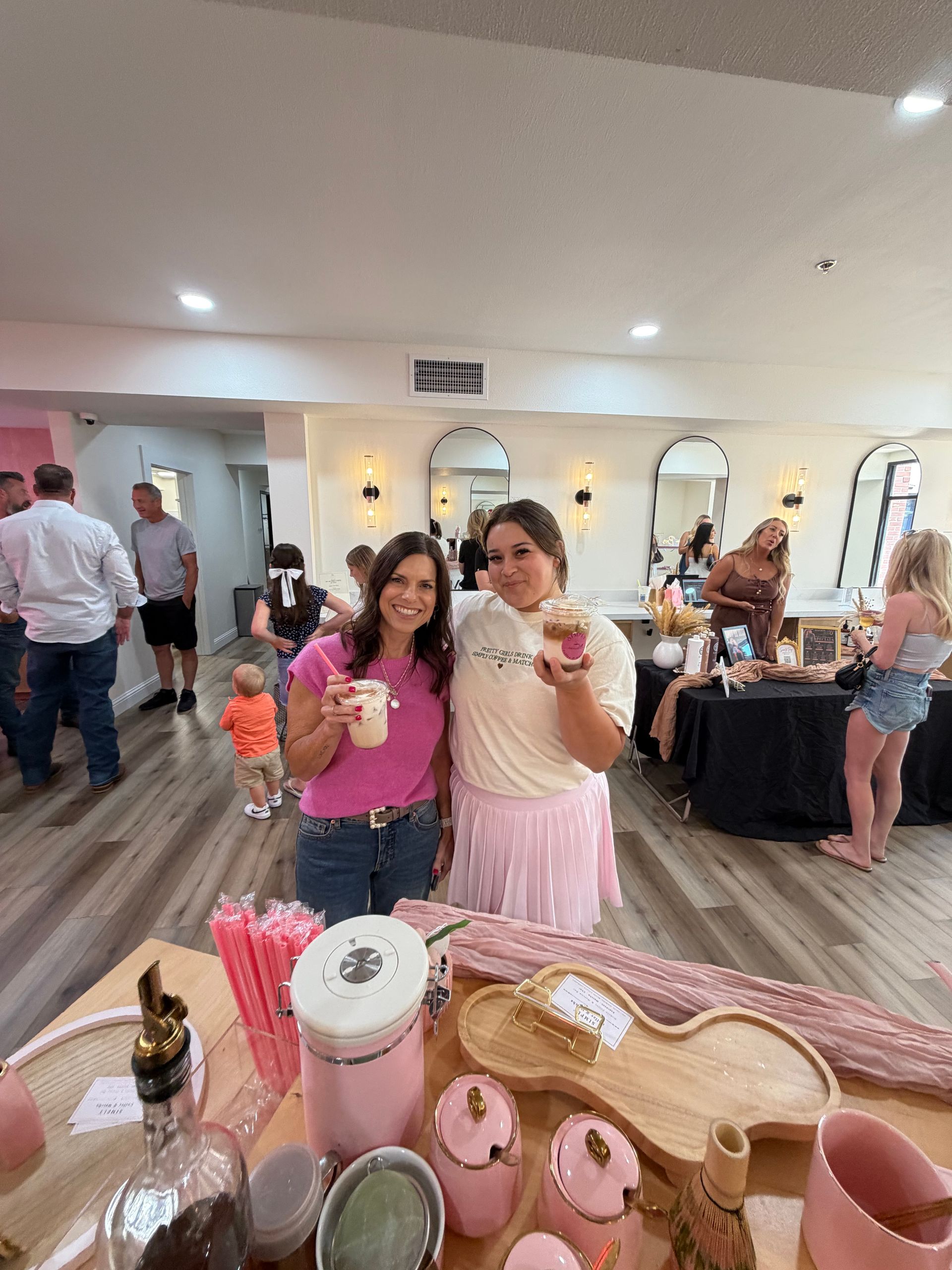 Two women in pink hold drinks at a lively indoor party with guests and a dessert table.