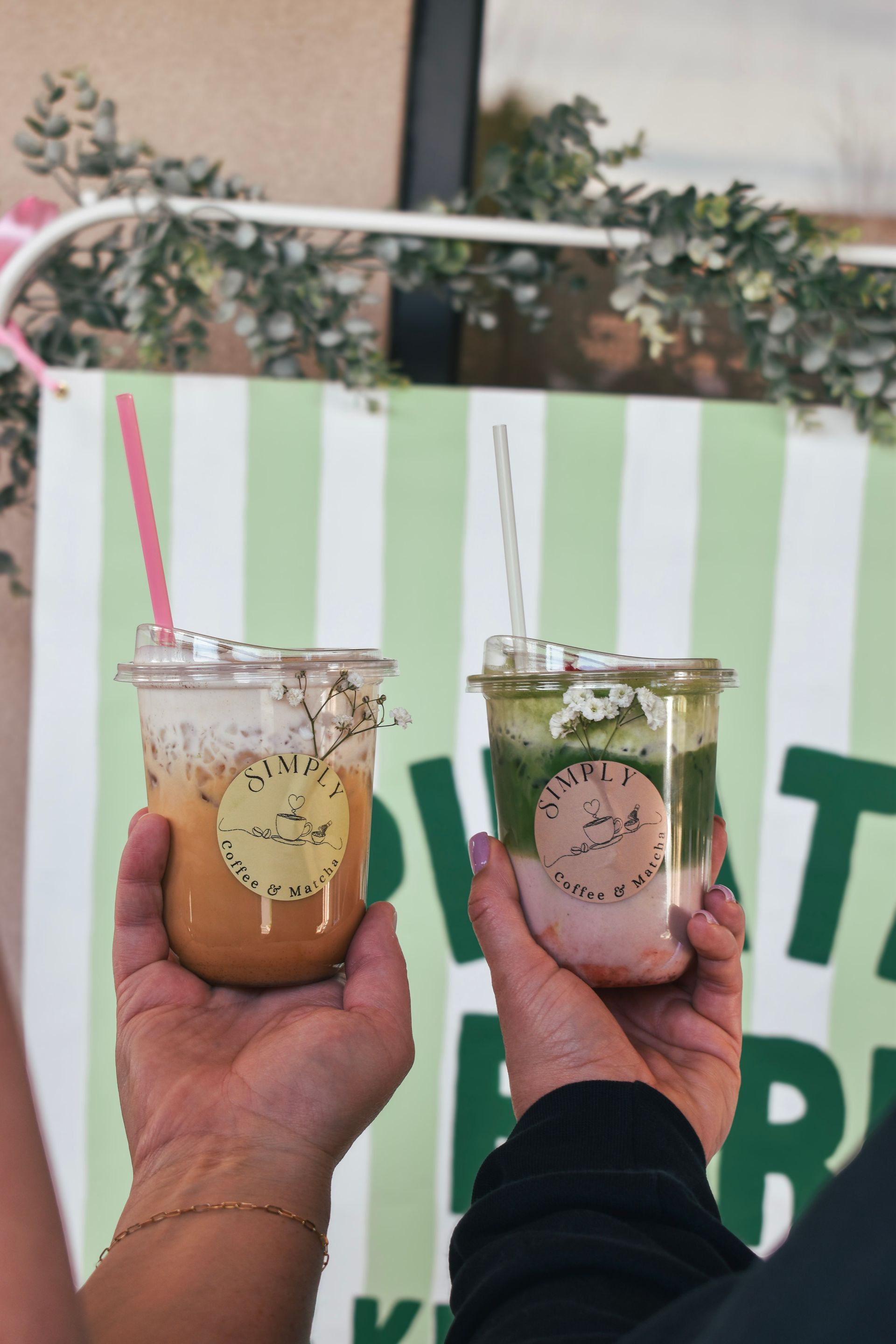 Two hands holding iced drinks with pink and green straws in front of a striped backdrop