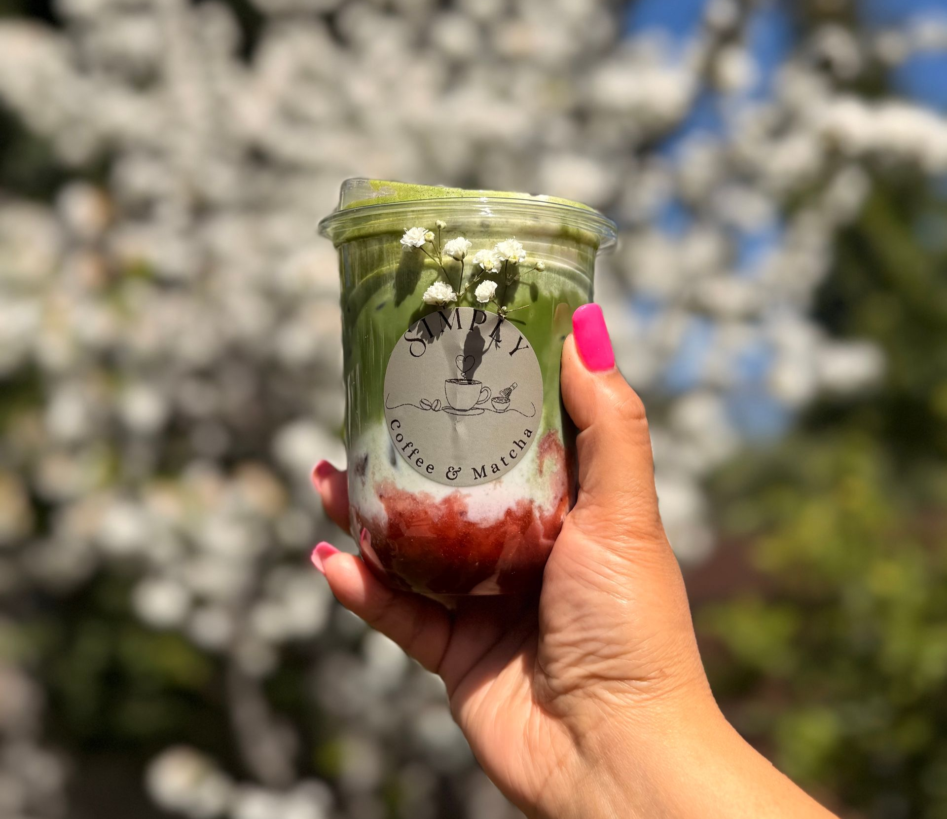 Hand holding a clear cup with green cream and red berry topping against a blurred floral background