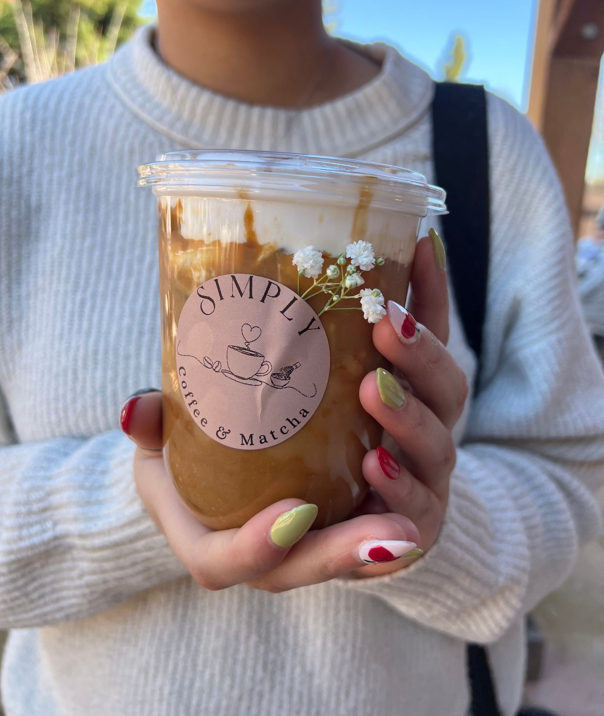Person holding a clear iced coffee cup with a pink logo, wearing a white sweater outdoors.