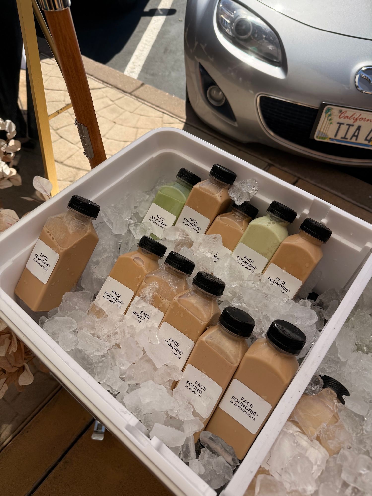 Assorted bottled drinks in an ice-filled cooler at an outdoor market, with a car parked behind it