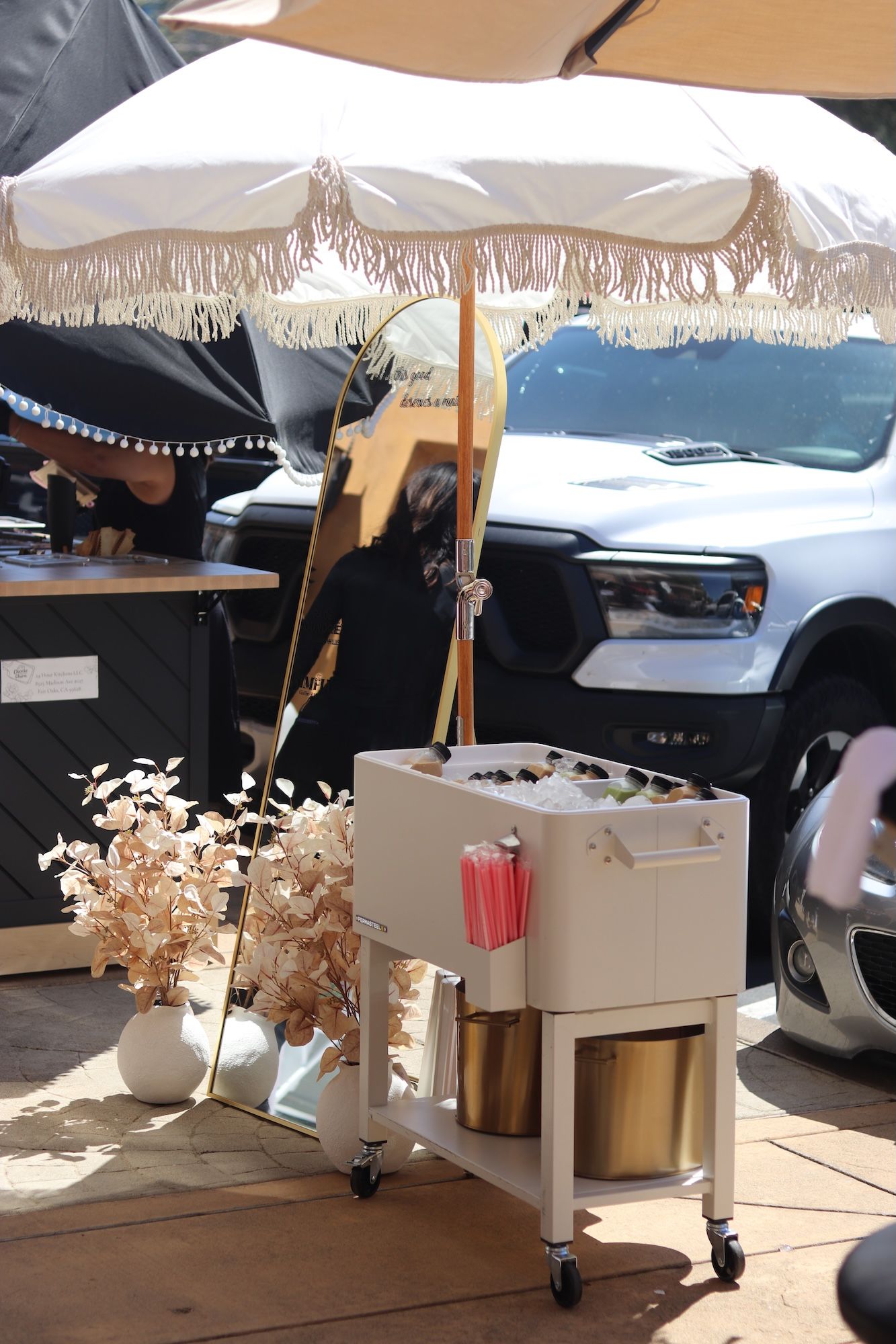 Outdoor coffee cart under a white umbrella beside a parked car, with cups and dried floral decor.
