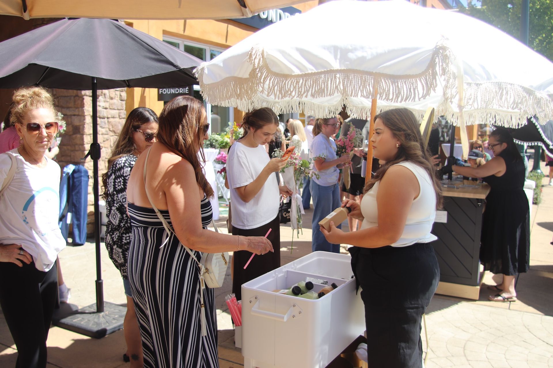 People mingling at an outdoor market, with umbrellas, a white cooler, and drinks being served