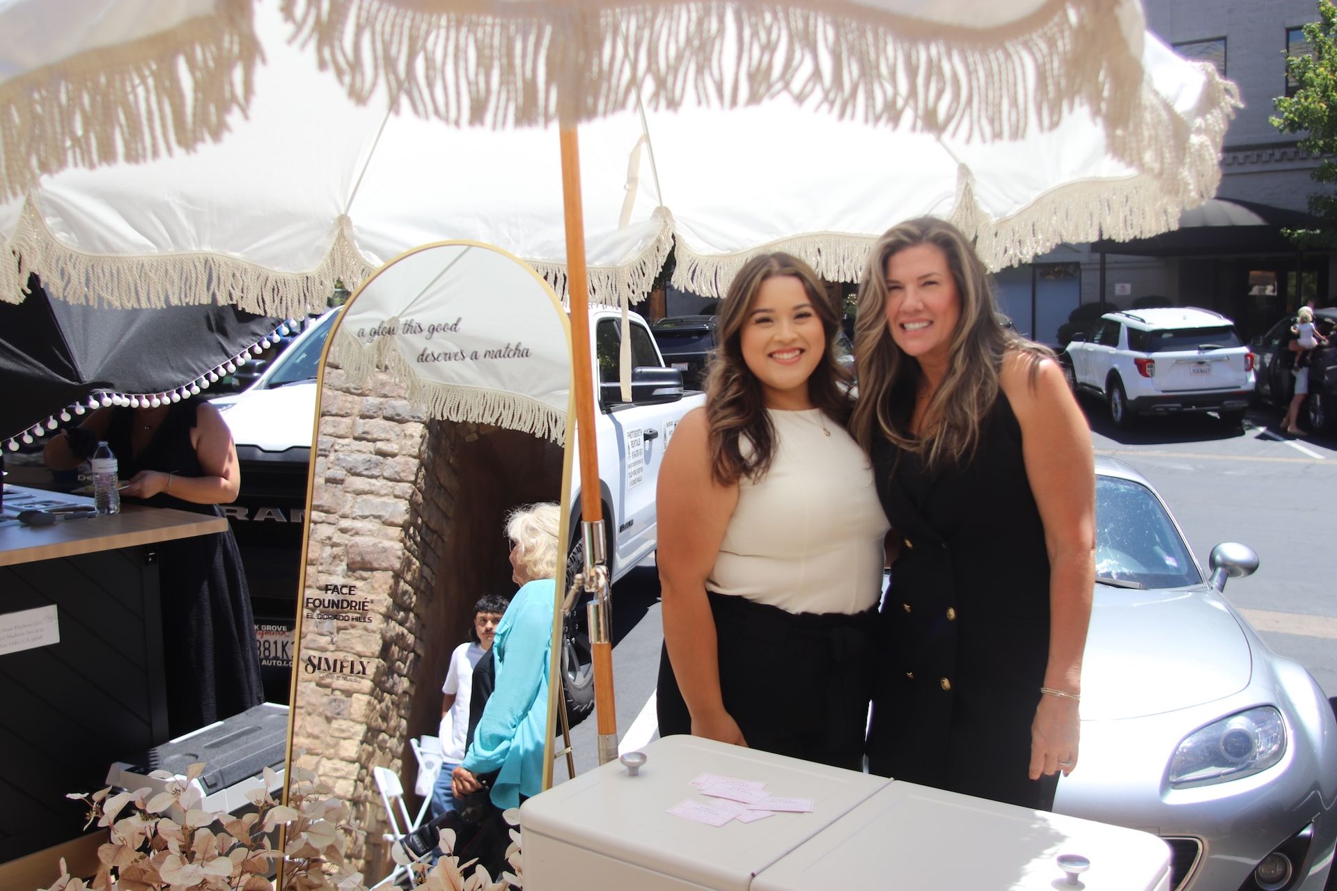 Two women smiling at an outdoor market stall beside a silver car under a white umbrella