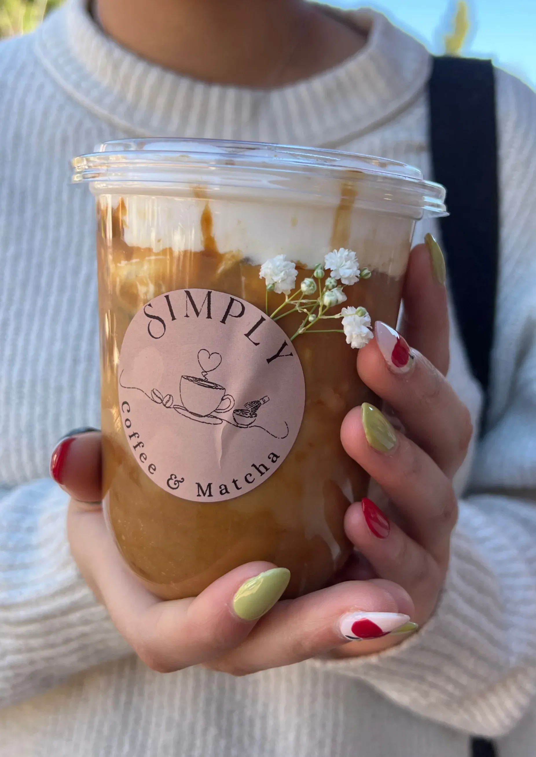 Hand holding iced coffee in a clear cup with pink logo, topped with whipped cream and flowers, pink nails visible