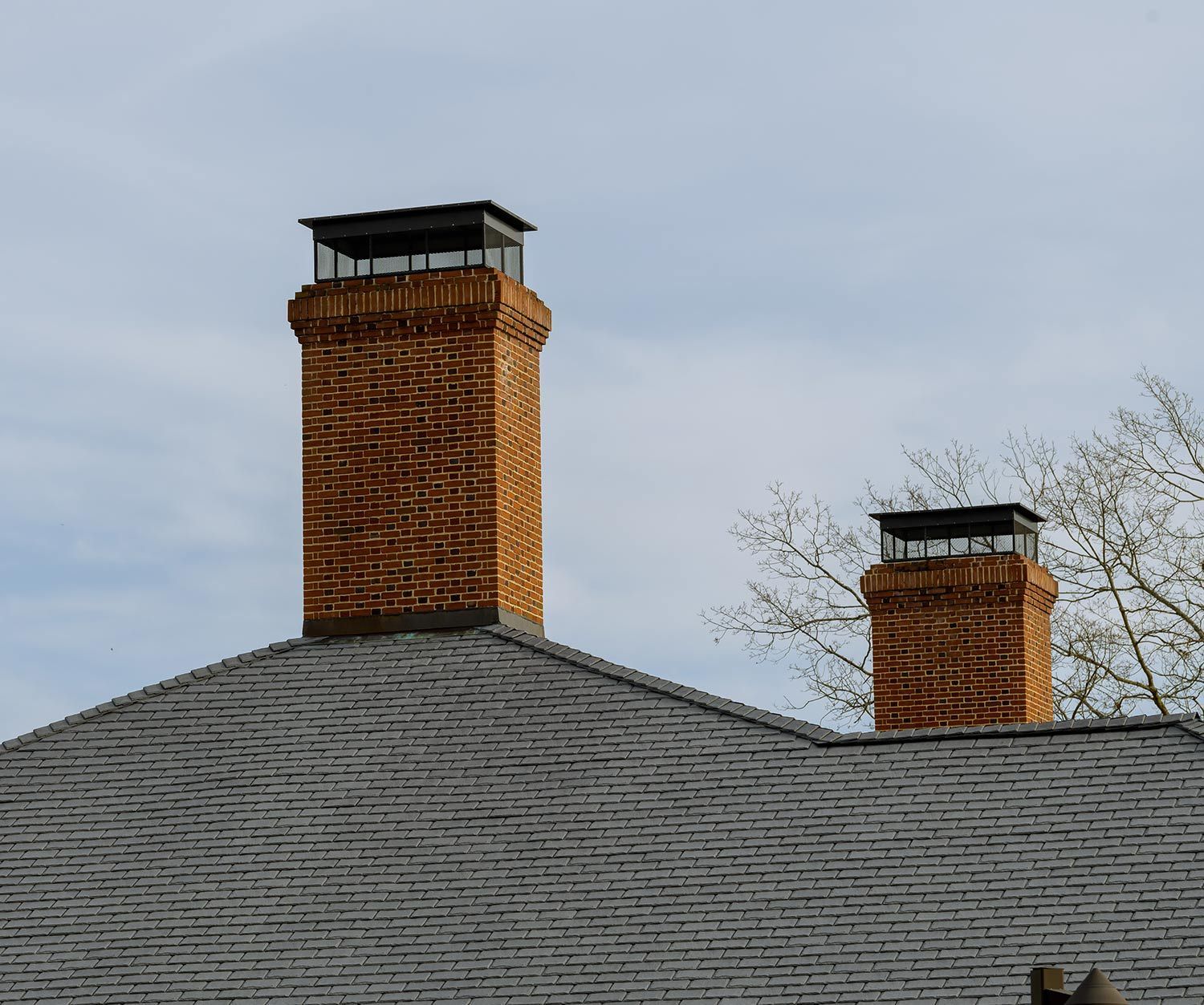 Two brick chimneys with black caps on a gray shingled roof under an overcast sky
