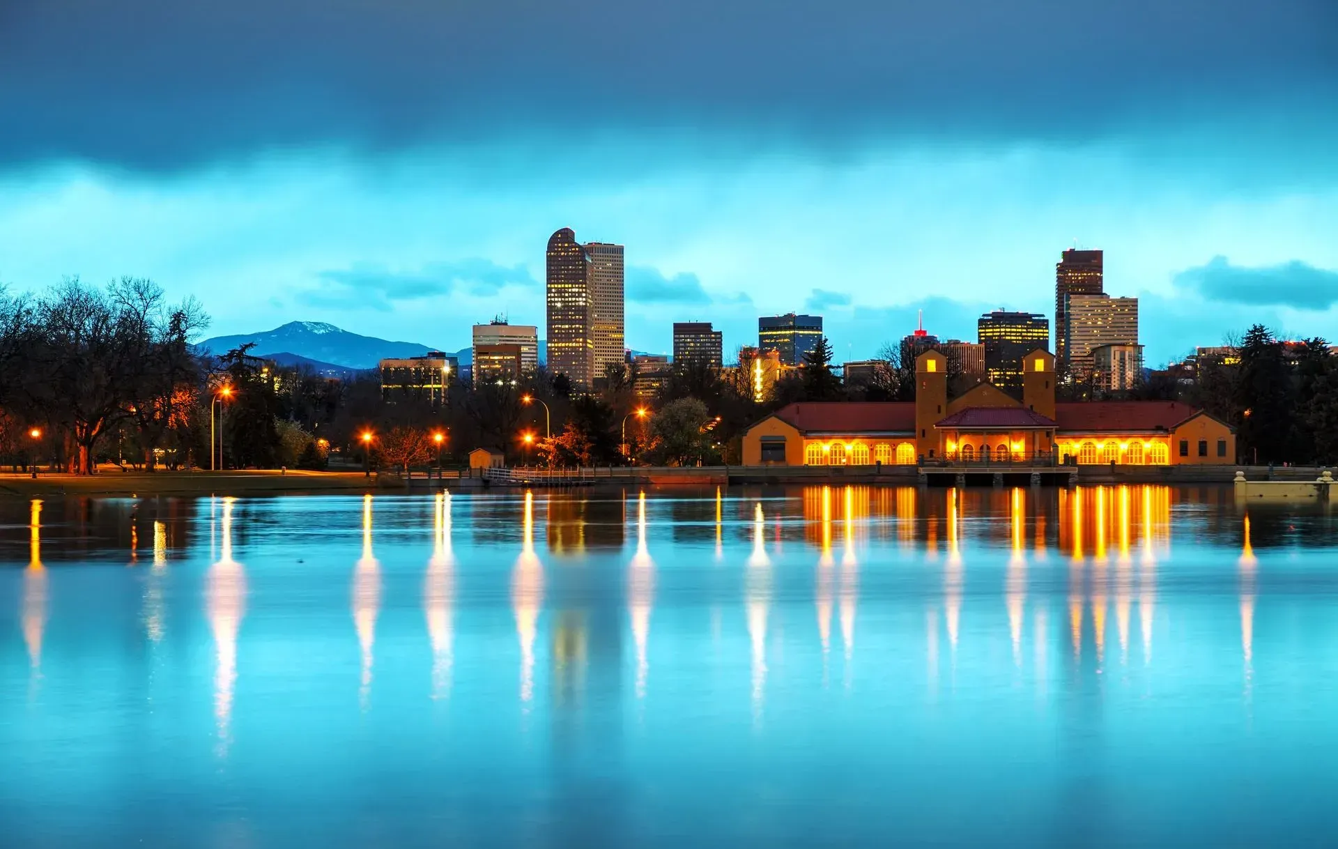 City skyline at dusk reflecting on a calm blue lake, with lit buildings and trees along the shore