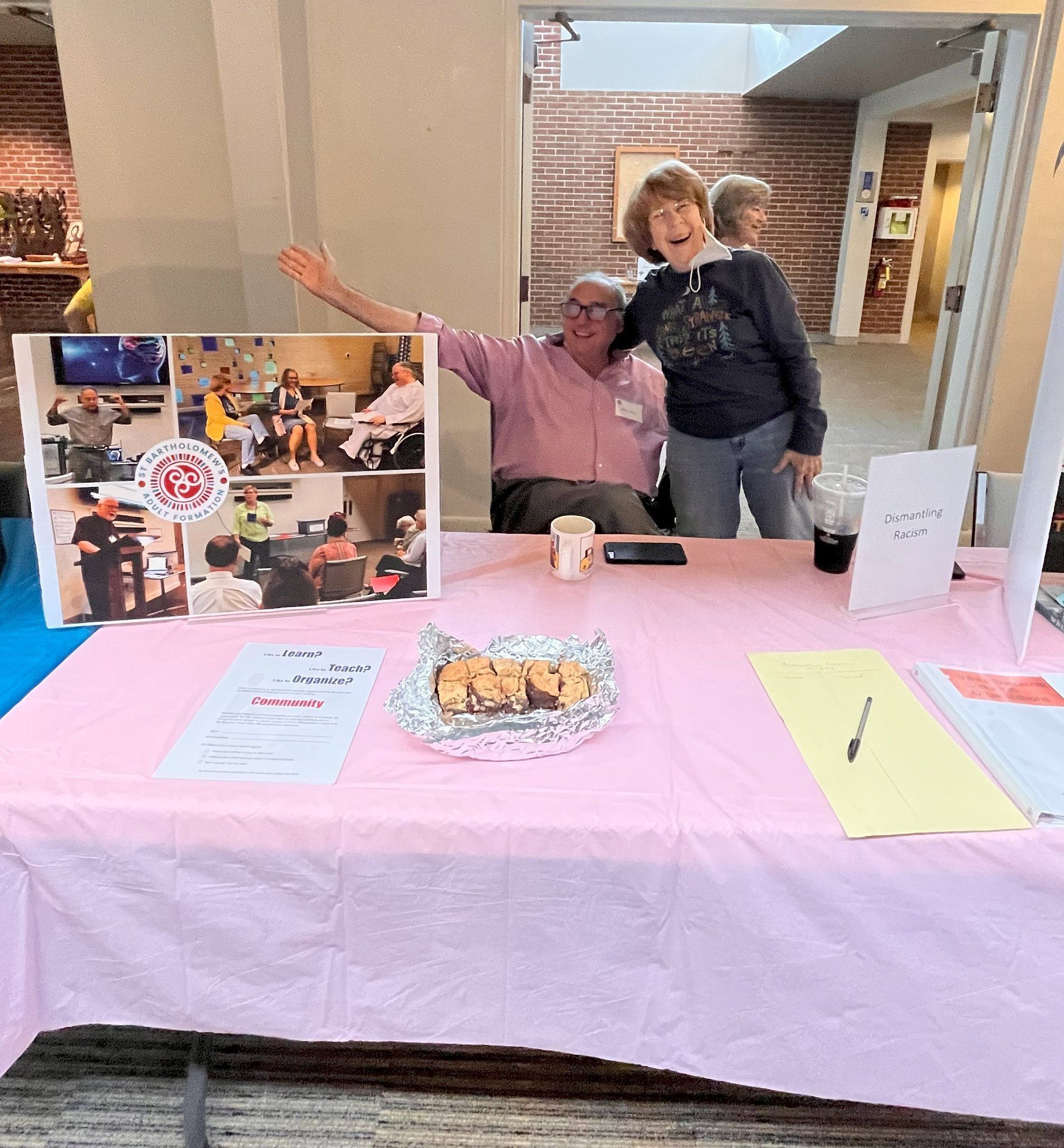 Two people behind a pink-covered table at a lobby display with posters and papers.