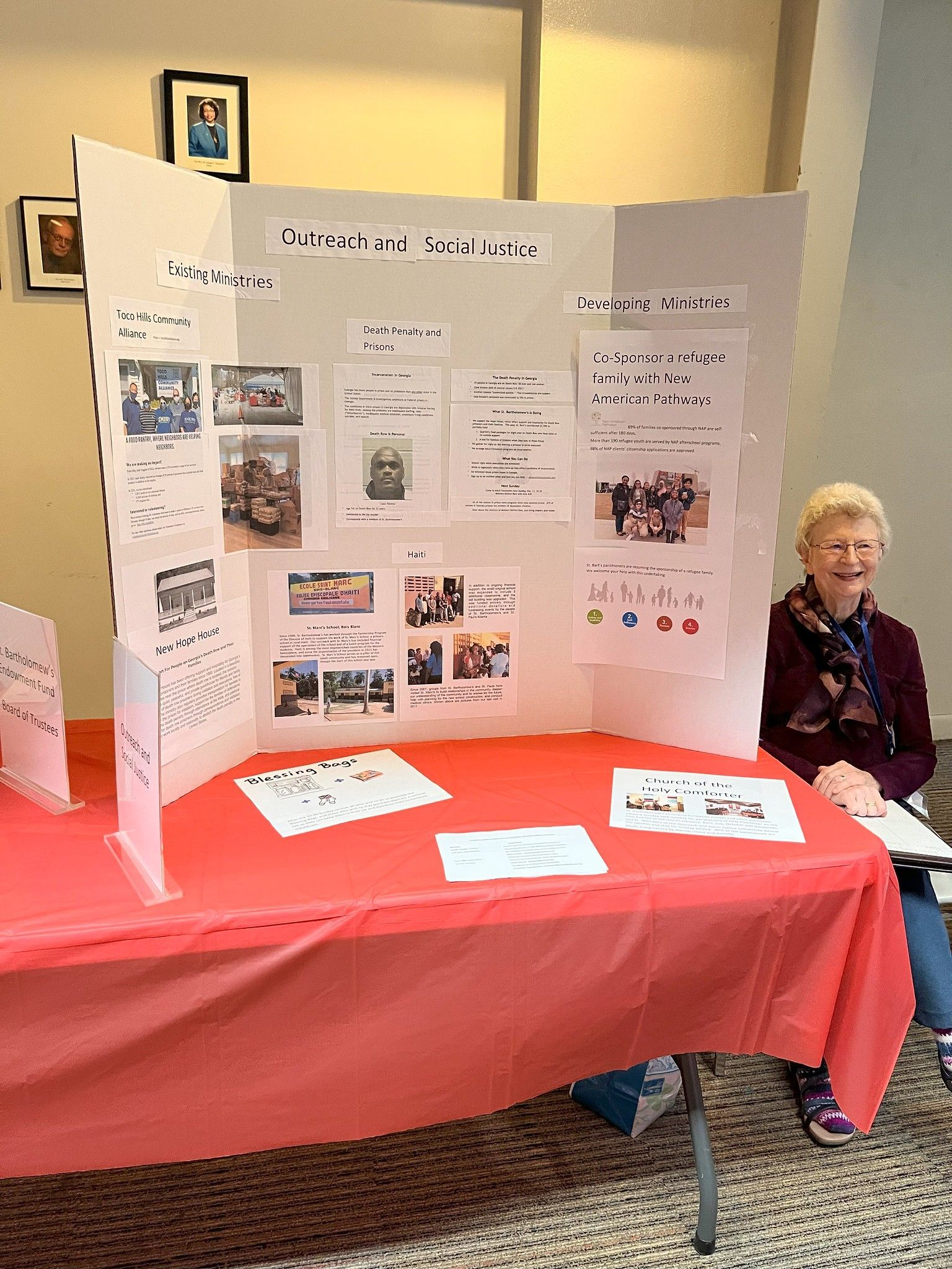 Display booth with tri-fold poster board on a red table, and a seated person beside it in a hallway.