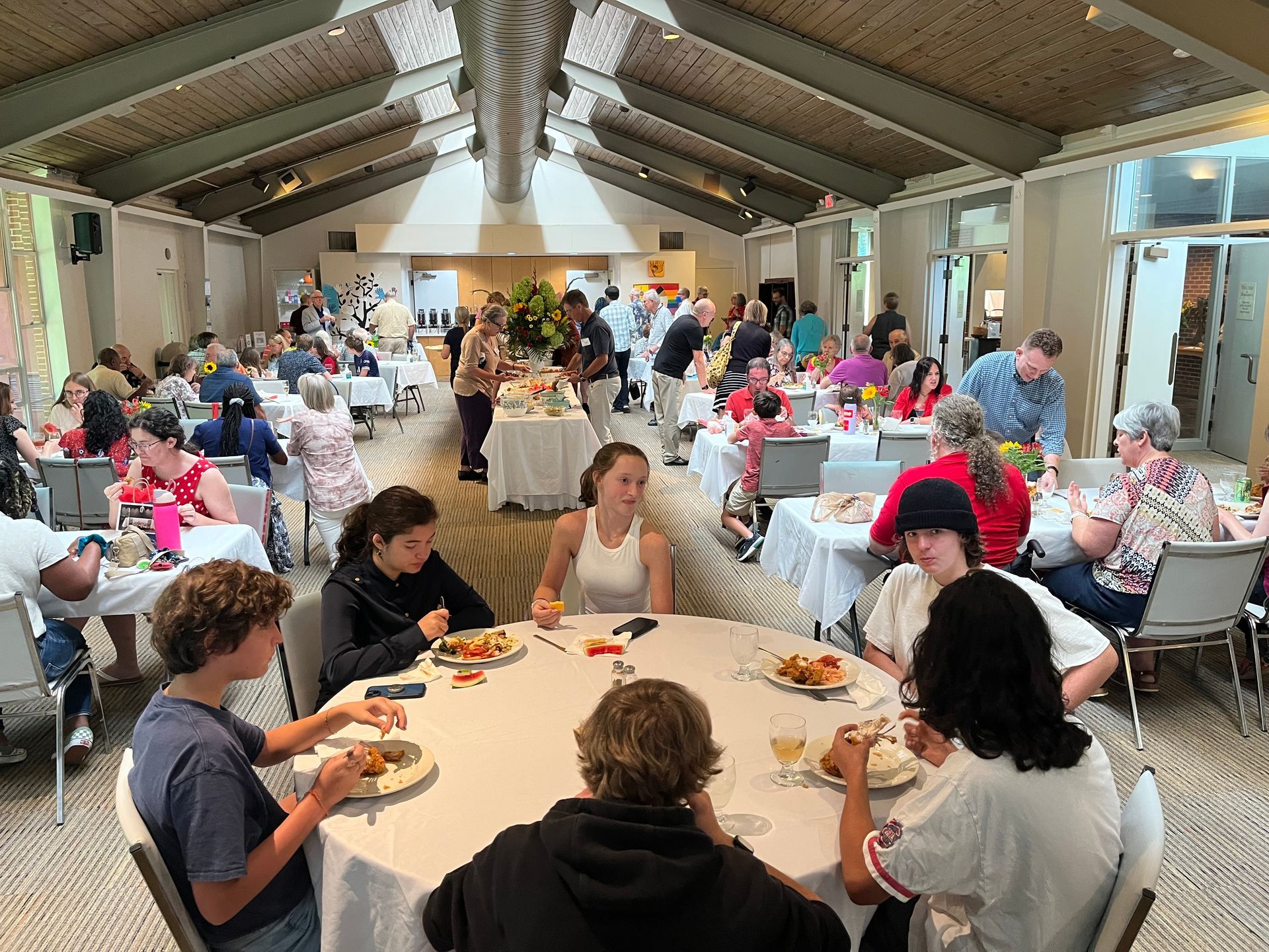 People seated at round tables in a bright banquet hall, eating and talking at a community gathering.