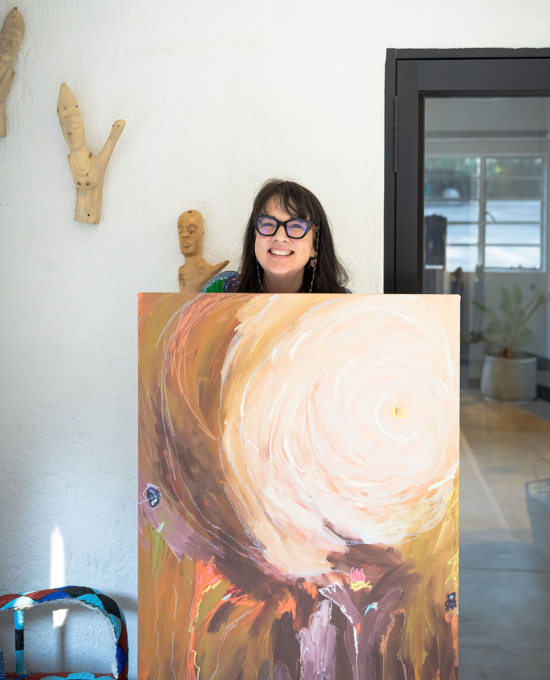 A person with dark hair and glasses smiles while holding a large, abstract painting with warm, earthy, circular colors.