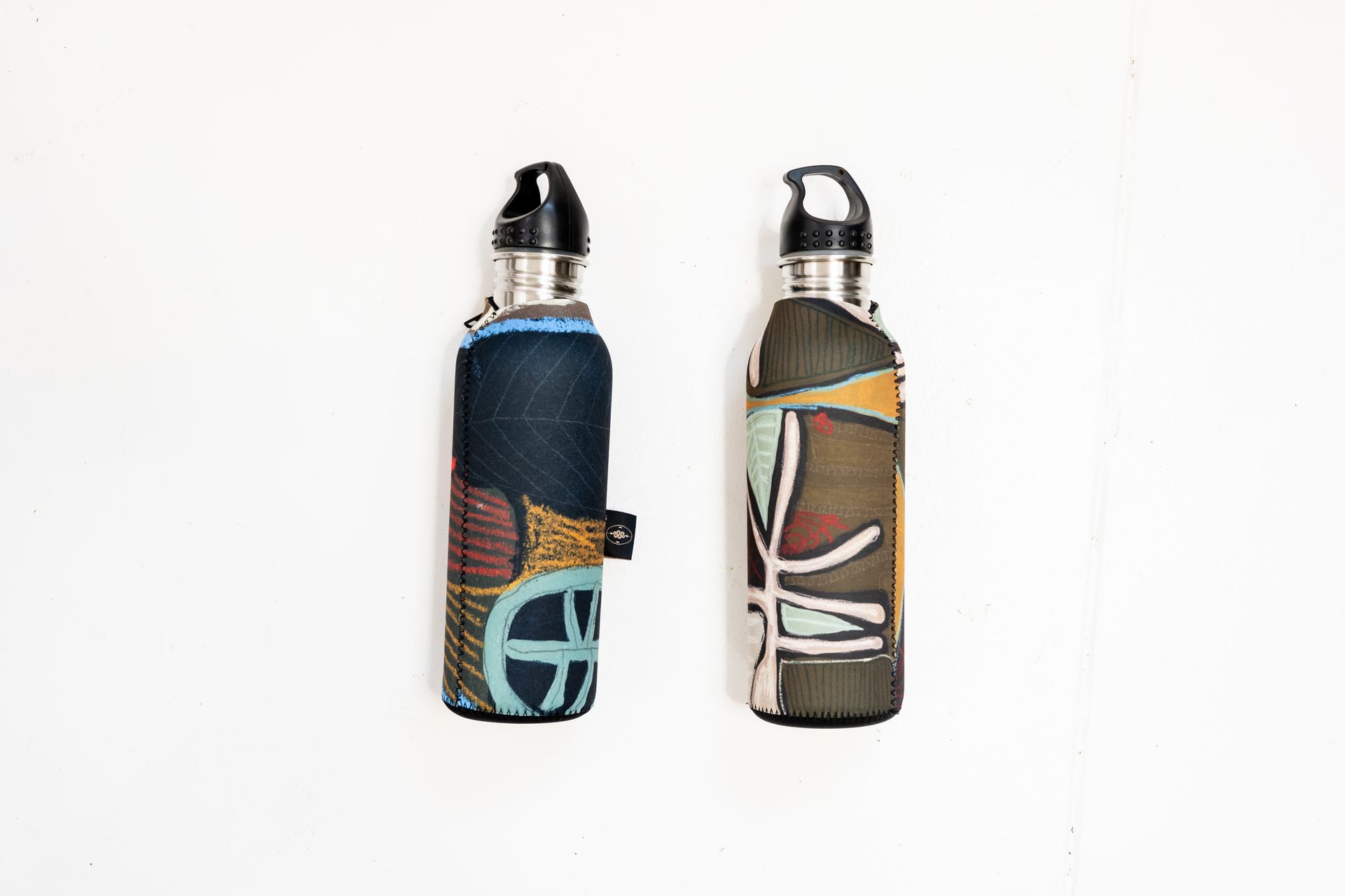 Two metal water bottles stand side-by-side, each encased in a patterned, fabric sleeve with a black carrying loop on top.