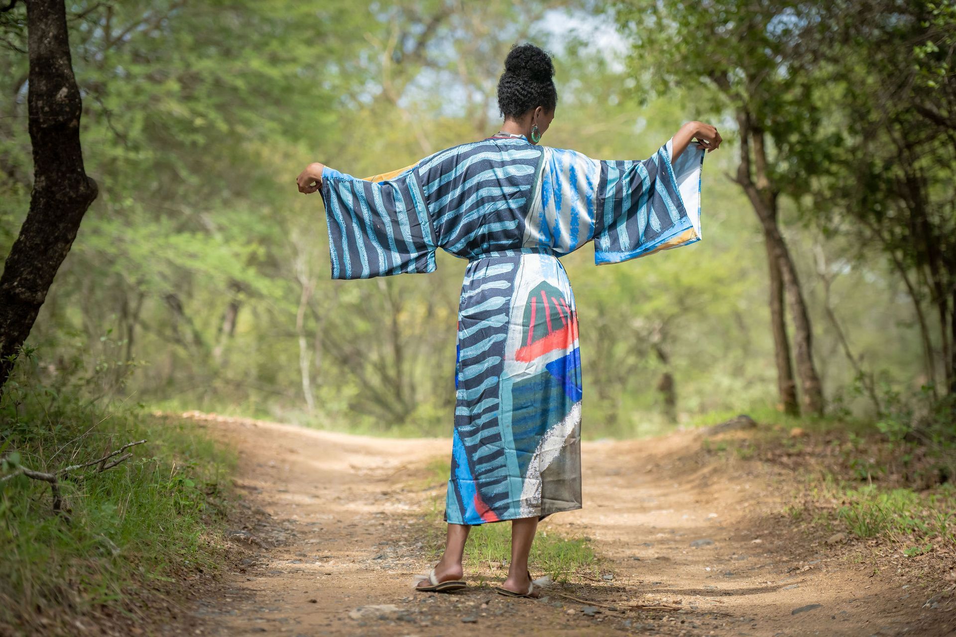 Person in a blue striped robe stands with arms out on a forest path.