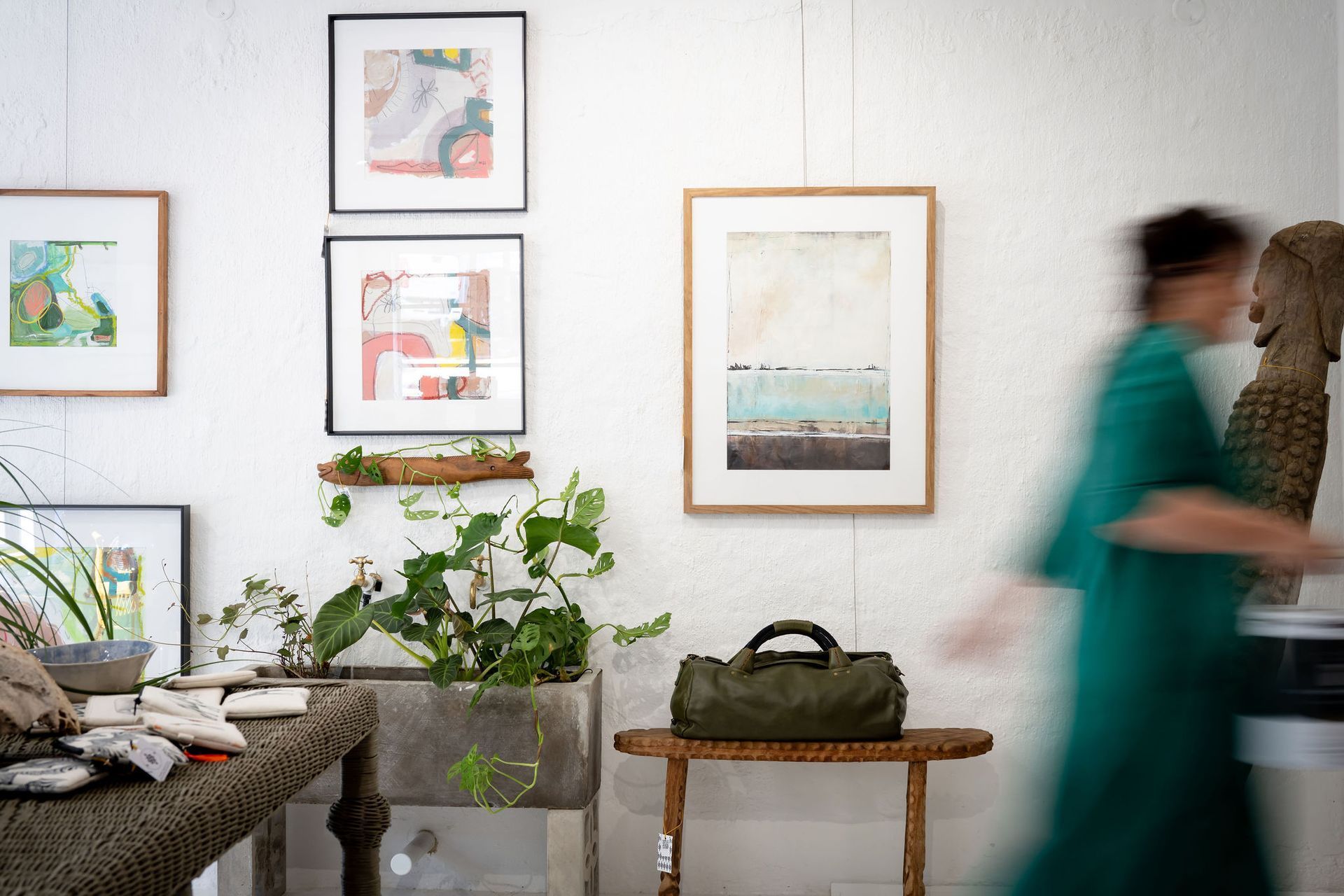 A blurry figure in green walks through a gallery space with framed art on white walls, a houseplant, and a leather bag.