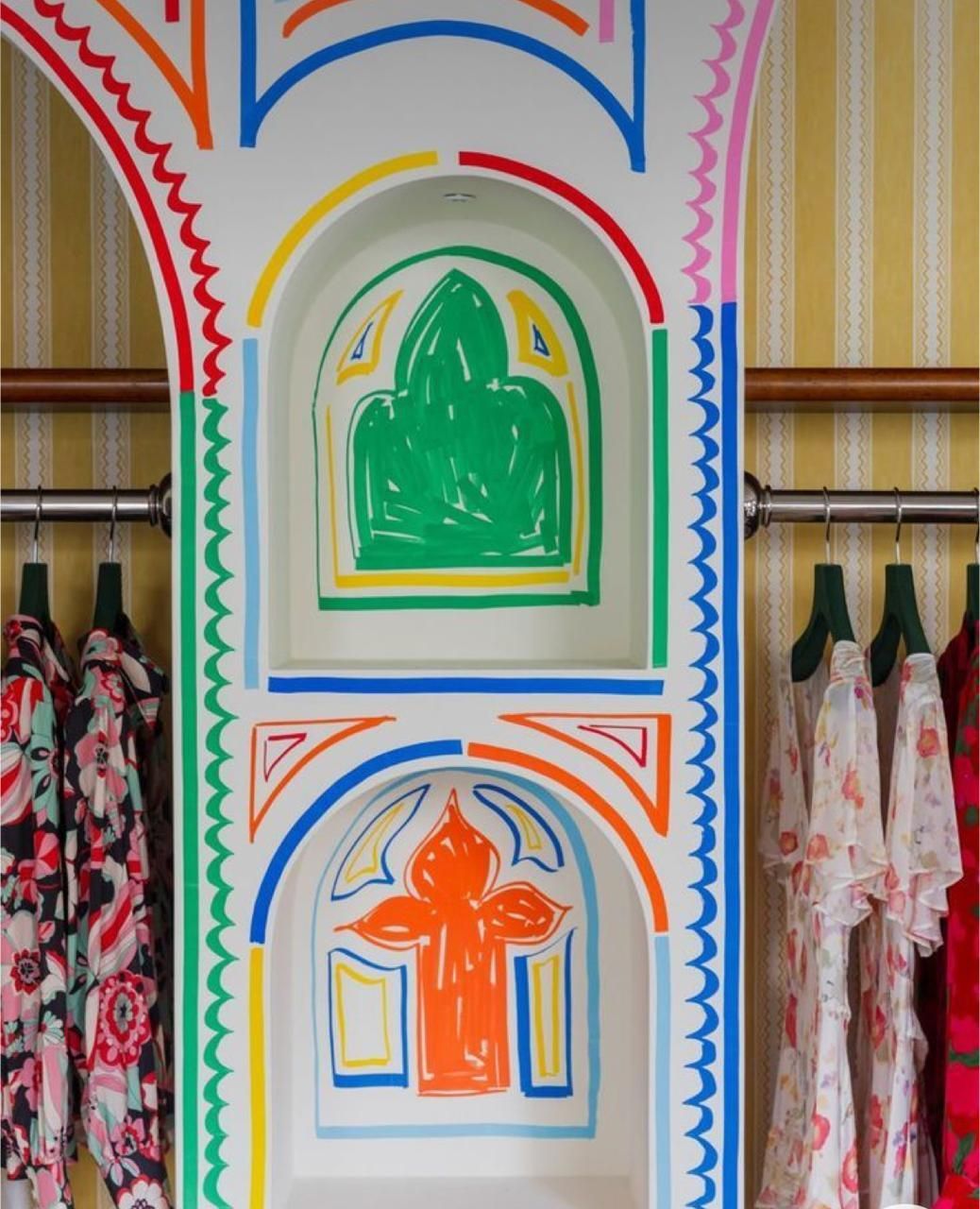 A central pillar painted with colorful, hand-drawn arches and shapes between clothing racks against striped wallpaper.