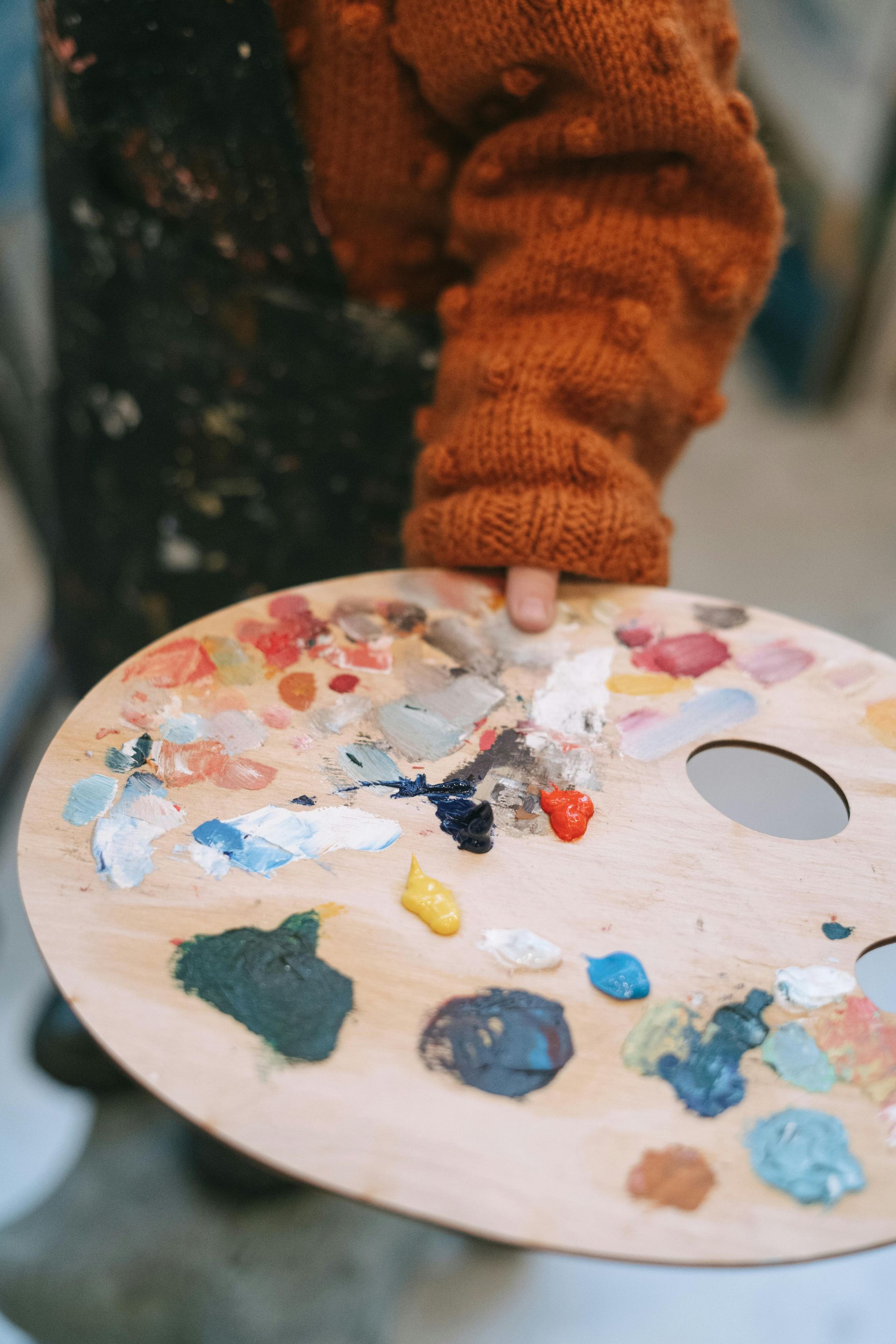 A person wearing a textured orange sweater holds a wooden artist's palette filled with various dabs of colorful paint.