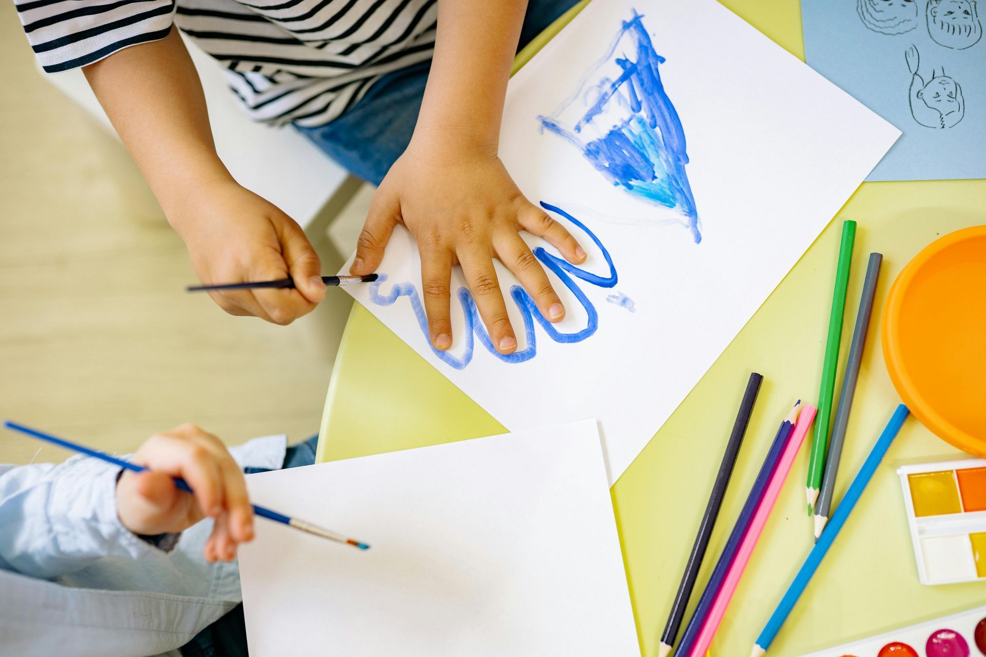 Two people paint on paper with brushes, with art supplies like pencils and paints on a table.