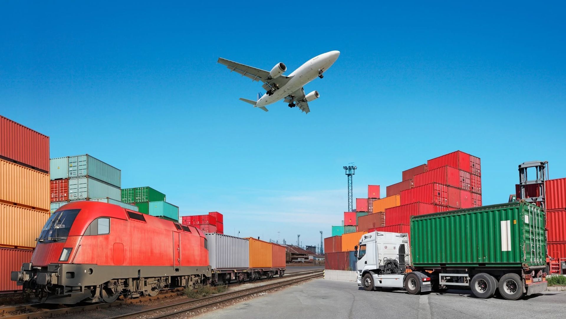 Cargo train yard with red and green freight containers and a plane flying overhead under a blue sky