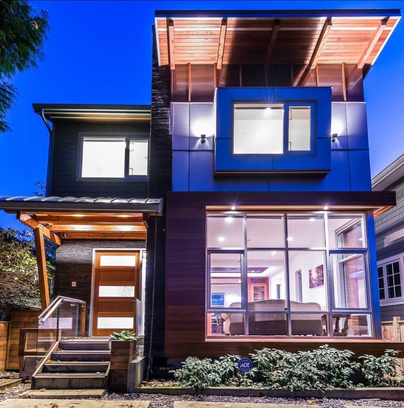 Modern two-story house exterior at dusk with large lit windows and a wooden front entry.
