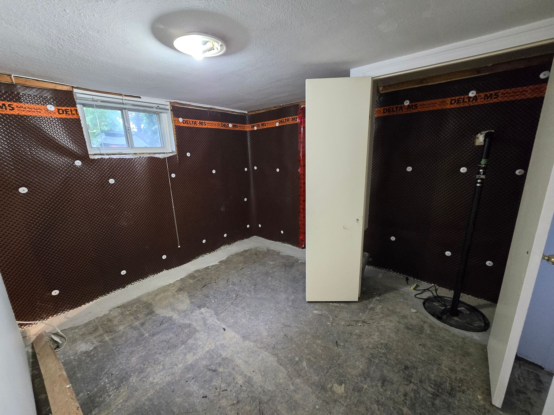 Basement interior with brown waterproofing membrane, small window, white cabinet, exposed concrete floor.